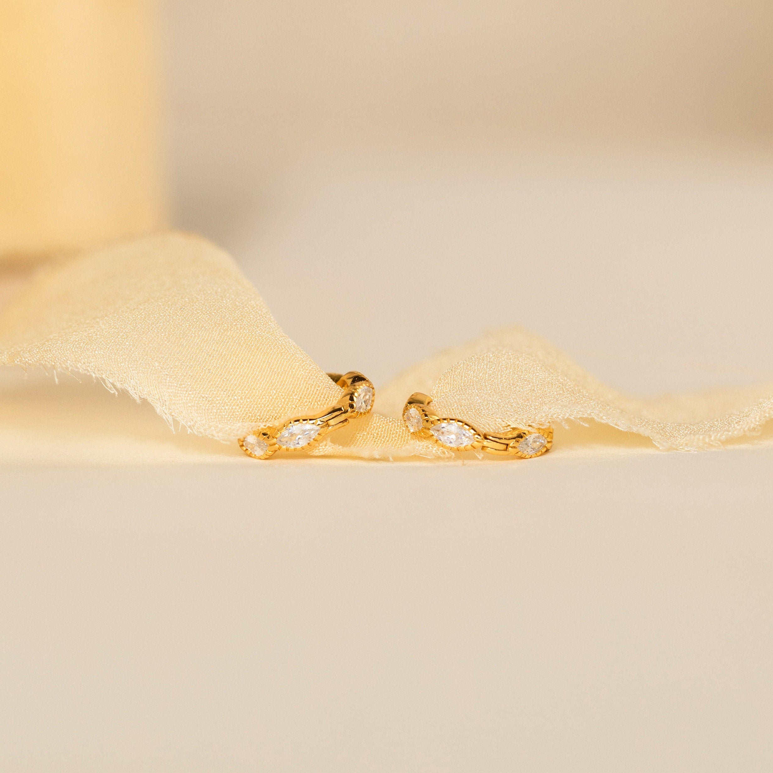 The Marquise Inlay Huggies—gold hoops with small, bezel-set gemstones—rest elegantly on light, sheer fabric.