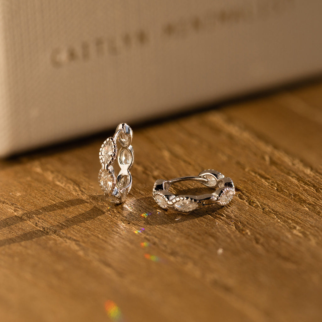 The Marquise Inlay Huggies, two silver hoops with intricate designs, are displayed on a wooden surface bathed in warm light.