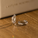 The Marquise Inlay Huggies, two silver hoops with intricate designs, are displayed on a wooden surface bathed in warm light.