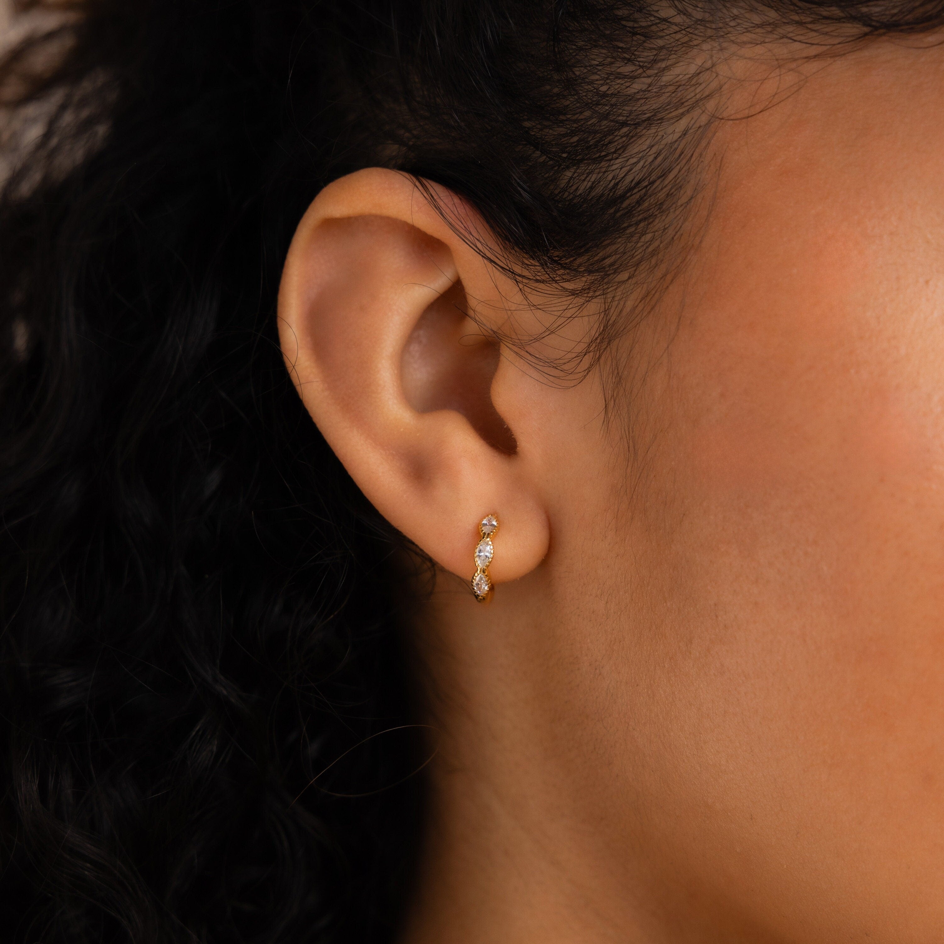 A close-up of a woman’s ear wearing Marquise Inlay Huggies—elegant gold huggie hoops adorned with three sparkling stones.