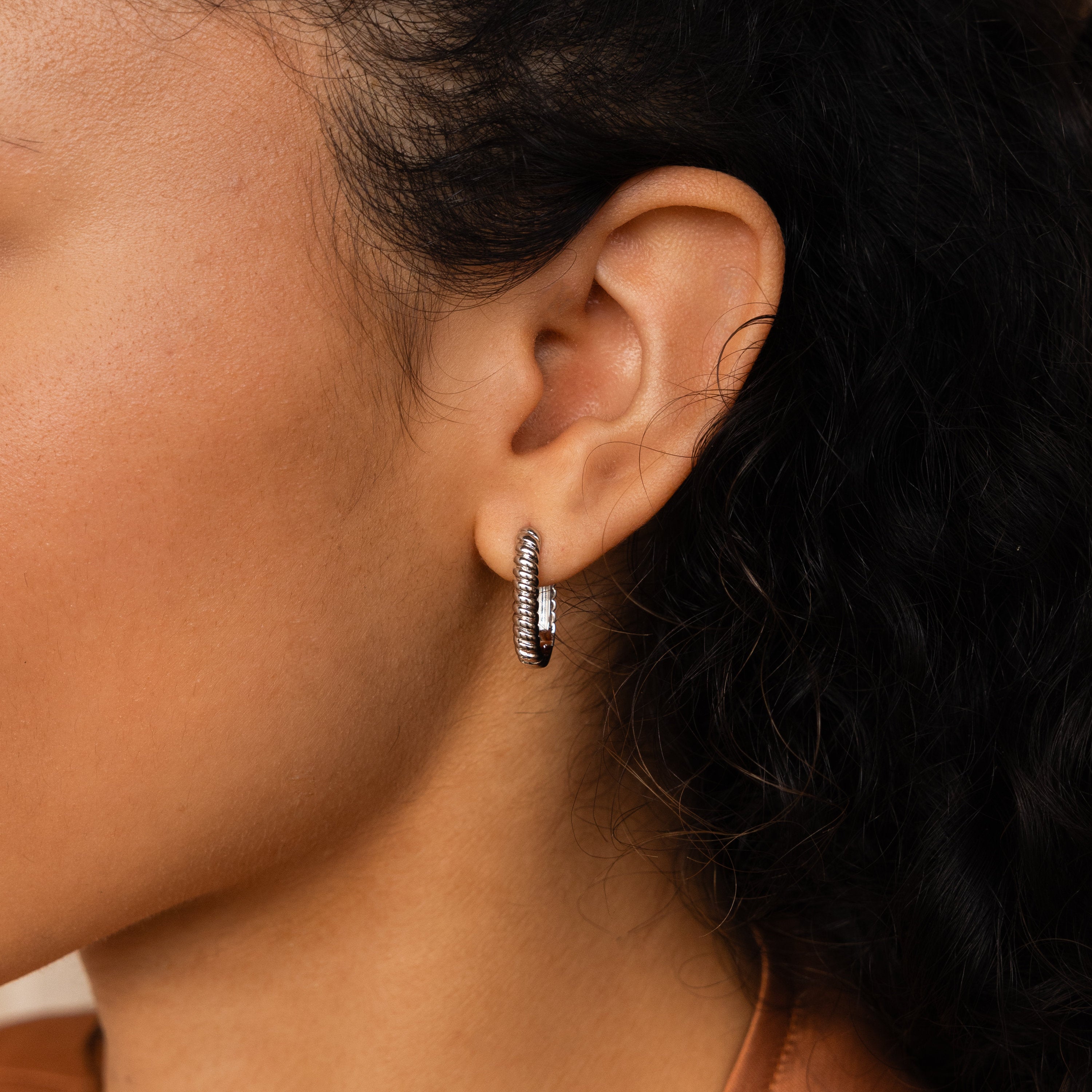 Close-up of a woman's ear wearing trendy Twisted Link Hoops with a ribbed texture, set against her curly dark hair.