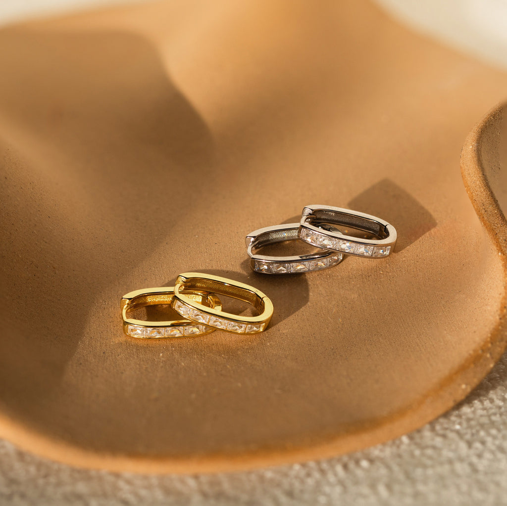 Two pairs of Baguette Link Hoops—one gold, one silver—adorned with embedded stones rest on a tan ceramic dish. Their chic, minimalist design makes them perfect for any occasion.