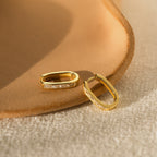 The Baguette Link Hoops, featuring gold with embedded stones and a geometric minimalist design, are displayed on a textured beige surface.