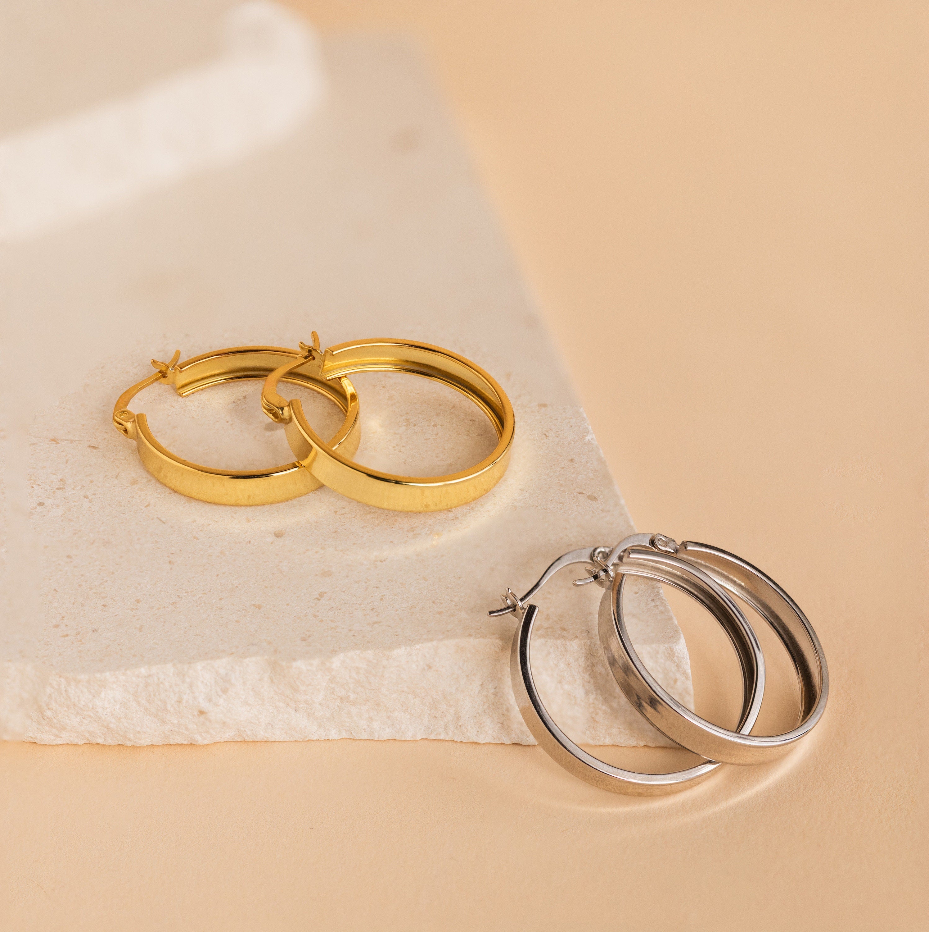 Thick Hoops in gold and silver are displayed on a textured beige stone slab against a neutral background, adding a bold, modern accent to any jewelry collection.