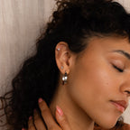 With closed eyes, a woman showcases our Thick Hoops silver earrings and her smooth skin as she gently touches her neck with a manicured hand.