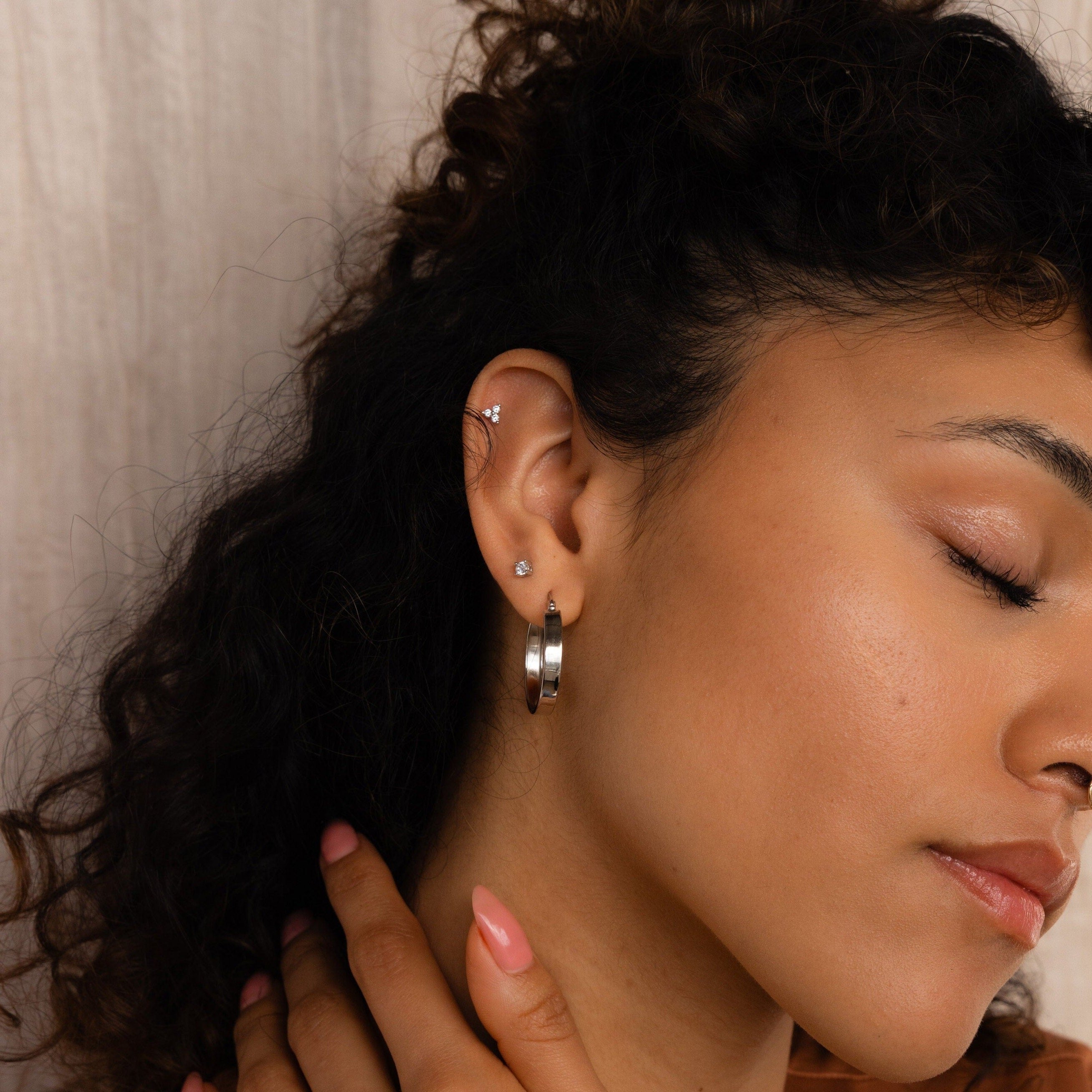 With closed eyes, a woman showcases our Thick Hoops silver earrings and her smooth skin as she gently touches her neck with a manicured hand.