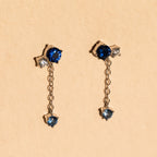 The Sapphire Diamond Drop Earrings feature blue and clear gemstones with a short dangling chain, elegantly displayed on a beige background.