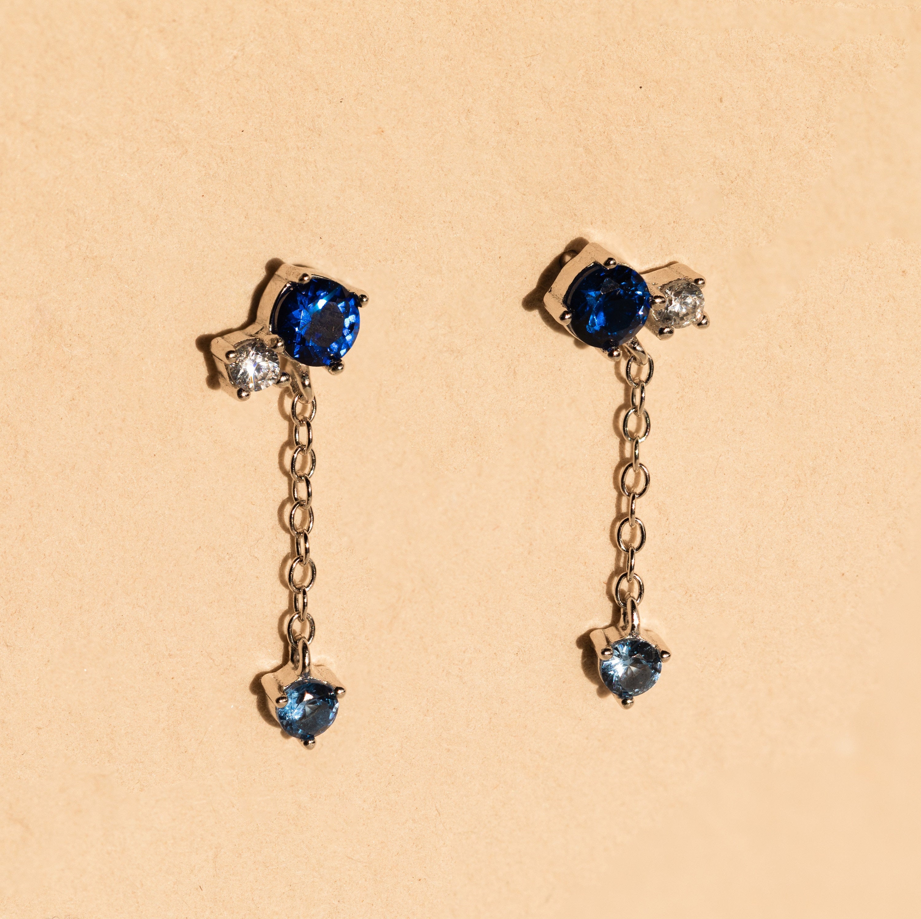 The Sapphire Diamond Drop Earrings feature blue and clear gemstones with a short dangling chain, elegantly displayed on a beige background.