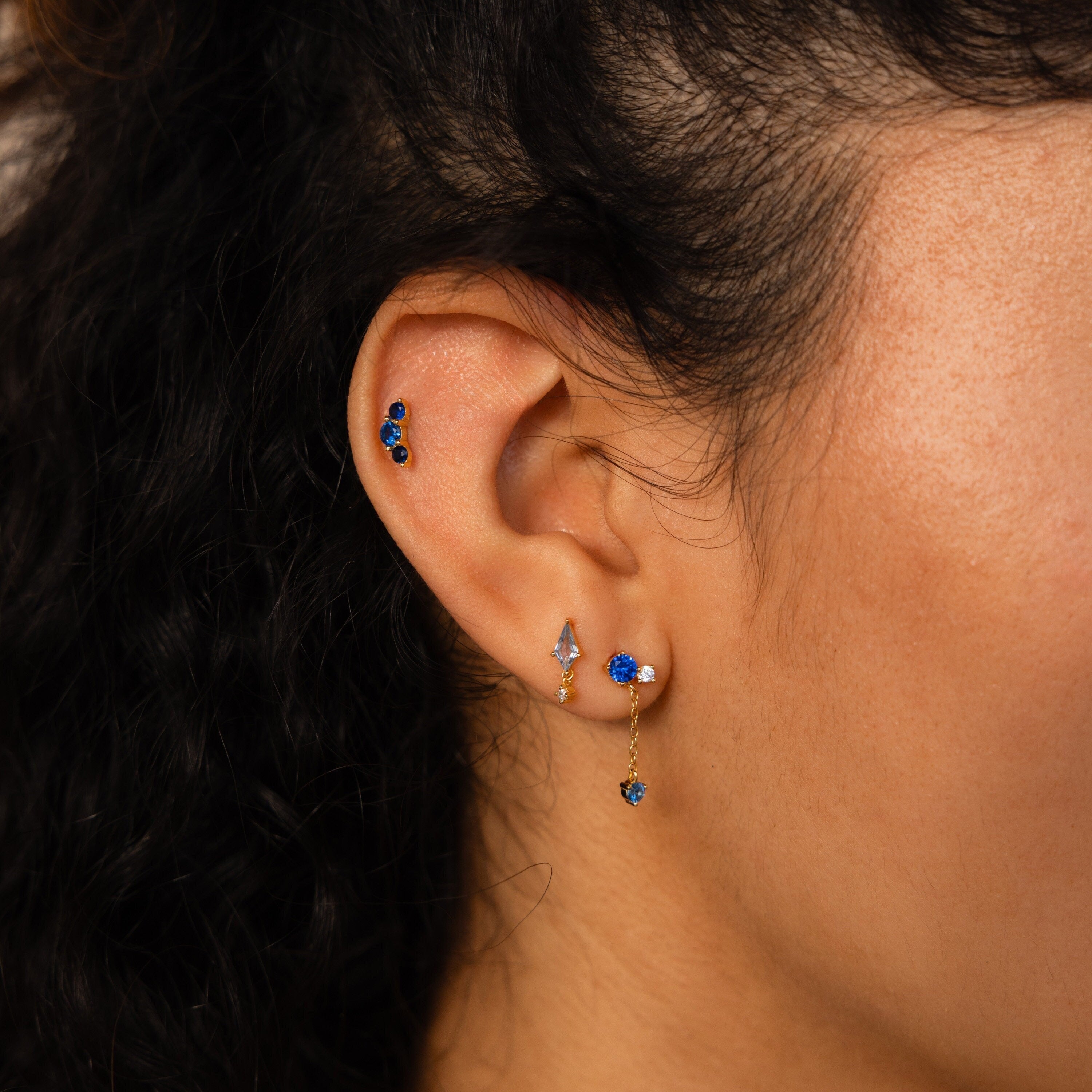 Close-up of a woman's ear with multiple gold earrings, featuring the Sapphire Diamond Drop Earrings and other sapphire and diamond pieces in various piercings.