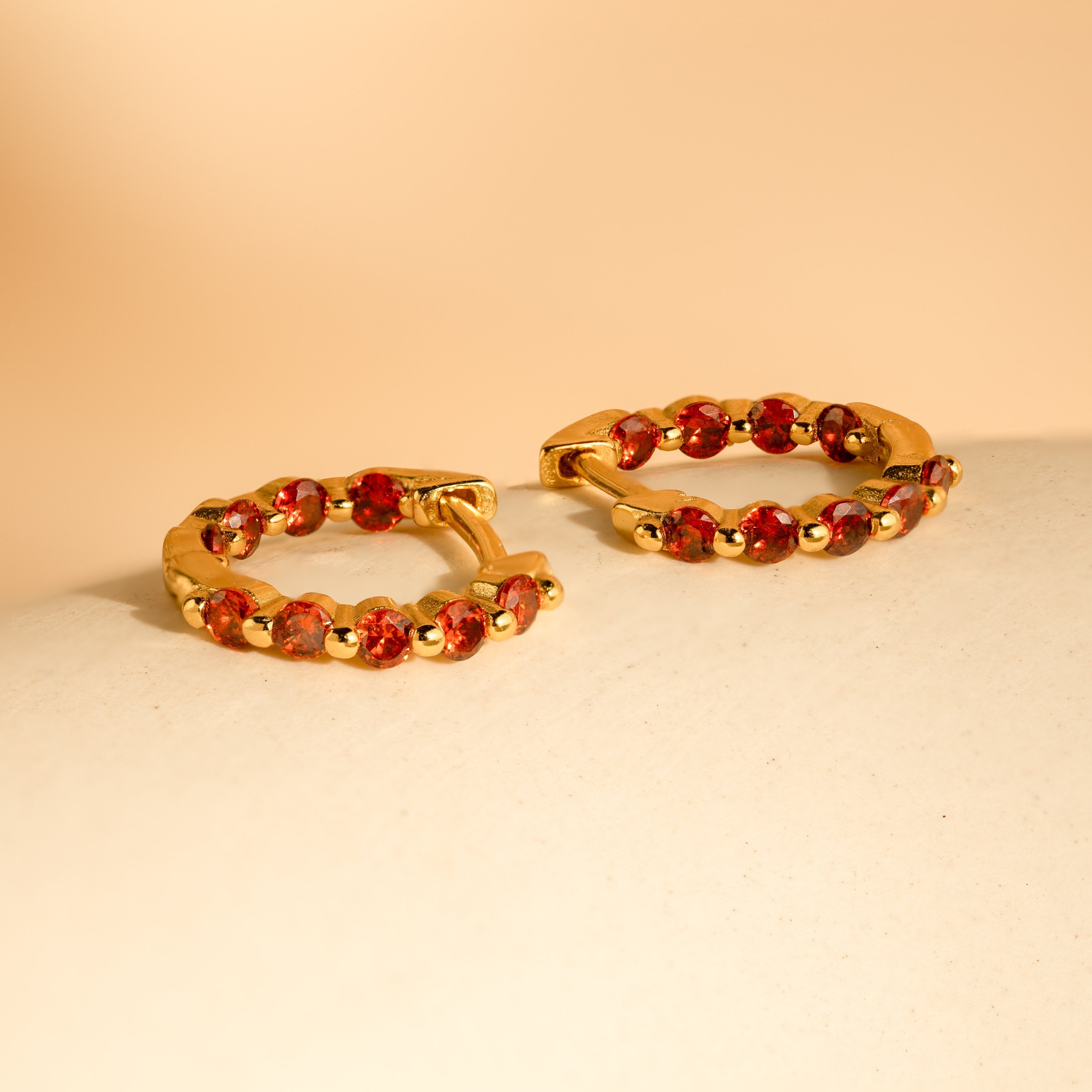 The Inside Out Huggies Set features gold hoop earrings with small red gemstones, displayed on a light beige surface—a perfect birthstone accessory to add a pop of color to your everyday look.