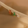 The Inside Out Huggies Set features three pairs of gold hoop earrings with vibrant red, green, and clear stones, creating a striking gemstone display on soft beige fabric.