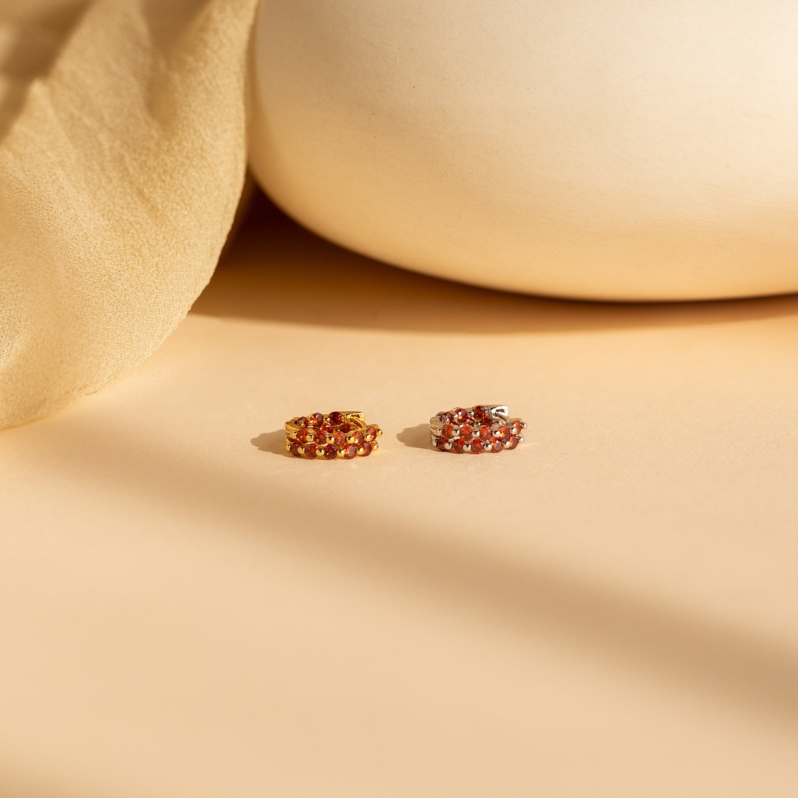 The Inside Out Garnet Huggies, featuring vibrant red and yellow gemstones, rest on a beige surface with soft fabric and pottery in the background.