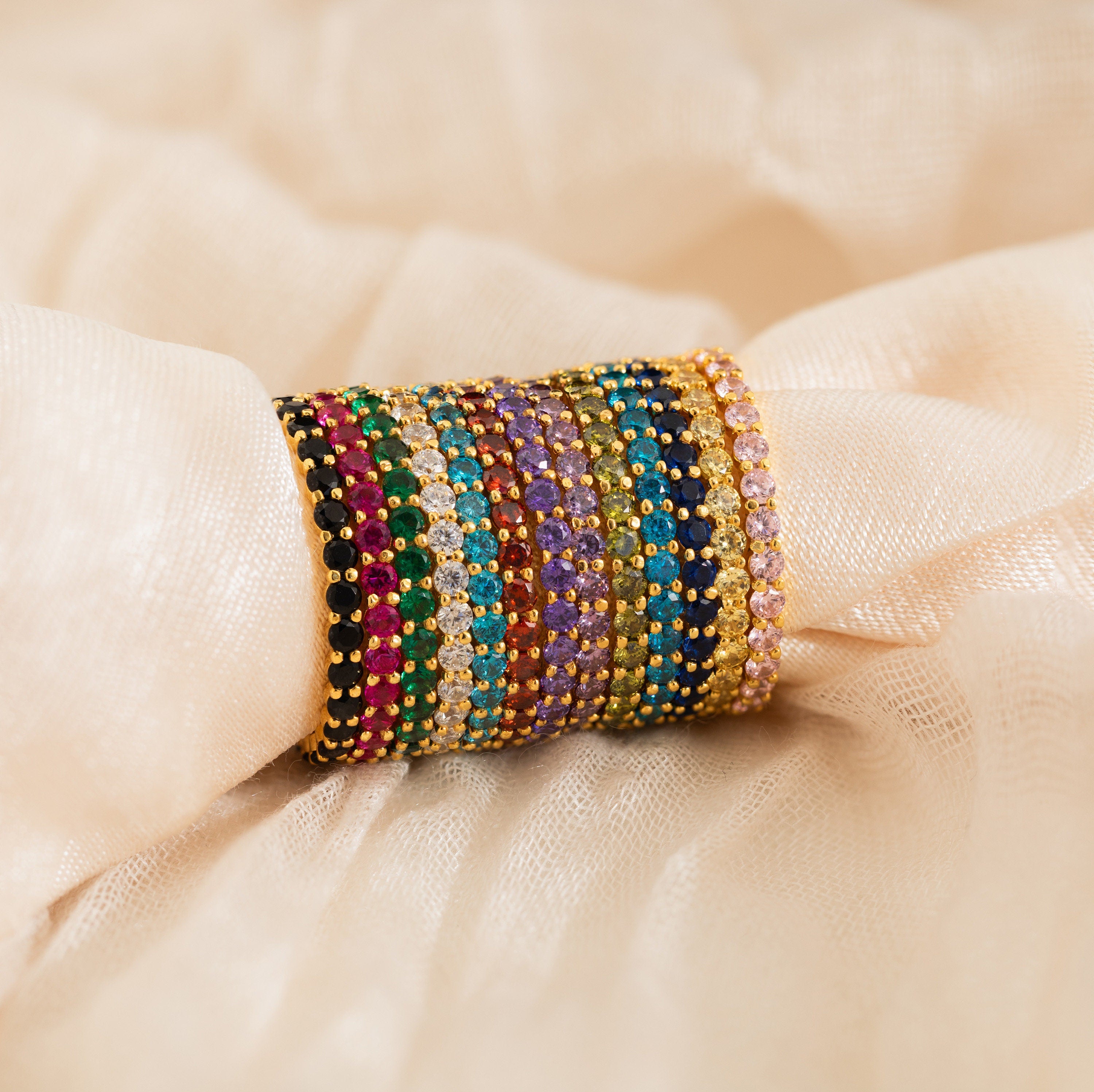 A stack of colorful gemstone-studded gold bangles rests on soft beige fabric, perfect as a Christmas gift or paired with the Eternity Tennis Birthstone Ring for added elegance.