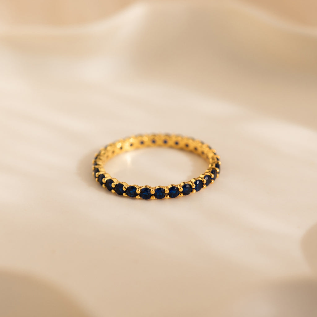 The Eternity Tennis Birthstone Ring, with small blue gemstones set all around, rests on a soft beige surface—a perfect customizable Christmas gift.