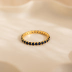 The Eternity Tennis Birthstone Ring, with small blue gemstones set all around, rests on a soft beige surface—a perfect customizable Christmas gift.