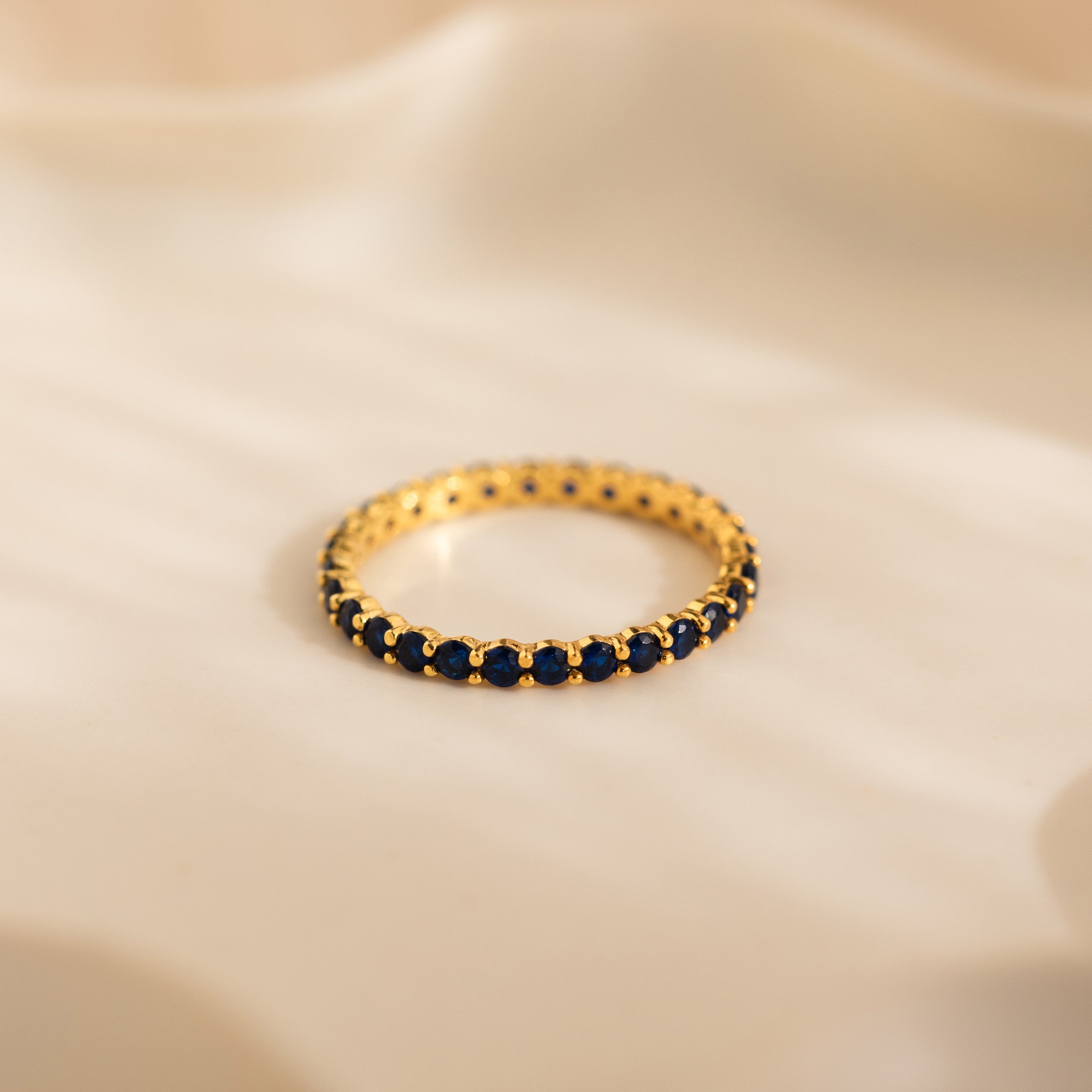 The Eternity Tennis Birthstone Ring, with small blue gemstones set all around, rests on a soft beige surface—a perfect customizable Christmas gift.
