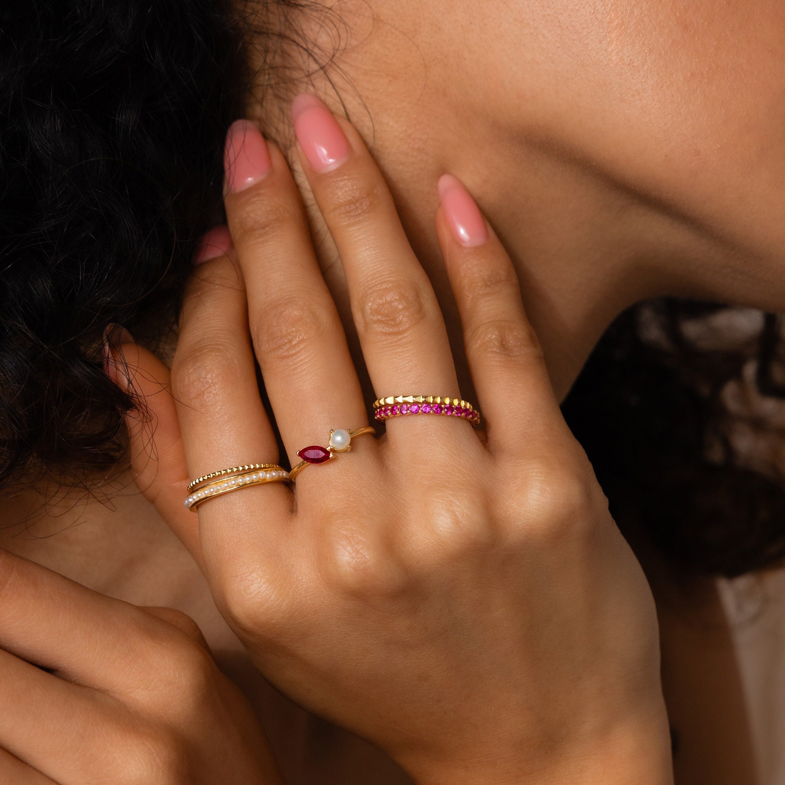 A hand with pink nails wears three gold rings, including the Eternity Tennis Birthstone Ring with diamonds, pearls, and pink gemstones, displayed near the face.