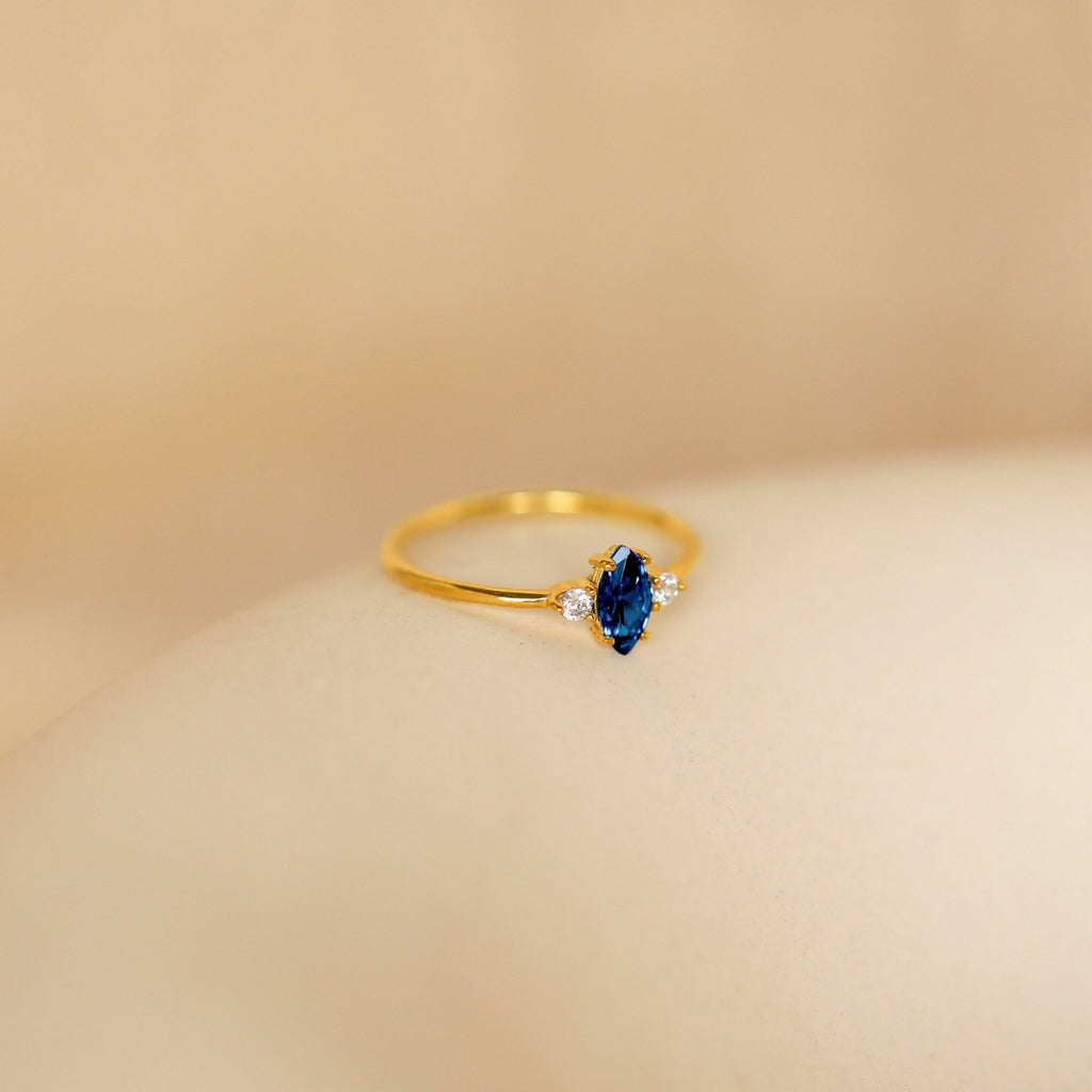 The Pave Marquise Birthstone Ring features a blue marquise gemstone with two small clear stones, ideal for custom birthstone jewelry, displayed on a soft beige background.