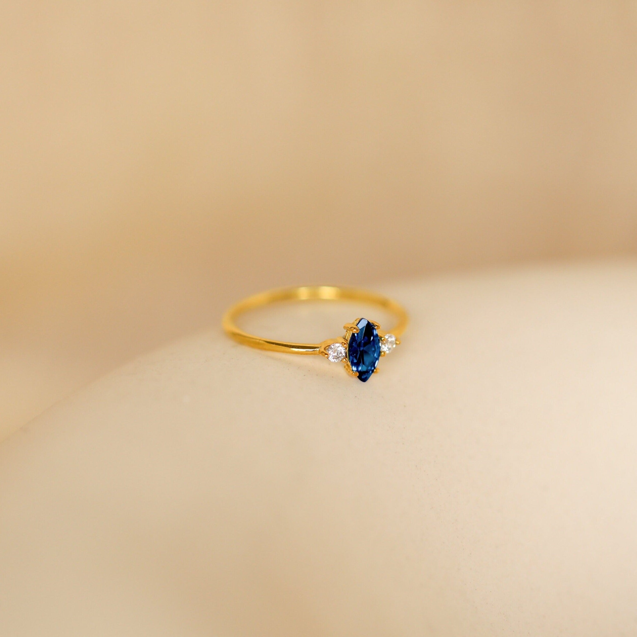The Pave Marquise Birthstone Ring features a blue marquise gemstone with two small clear stones, ideal for custom birthstone jewelry, displayed on a soft beige background.