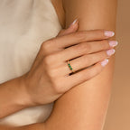 A woman's hand wearing the Pave Triple Birthstone Ring with green gemstones rests on her arm against a beige background.