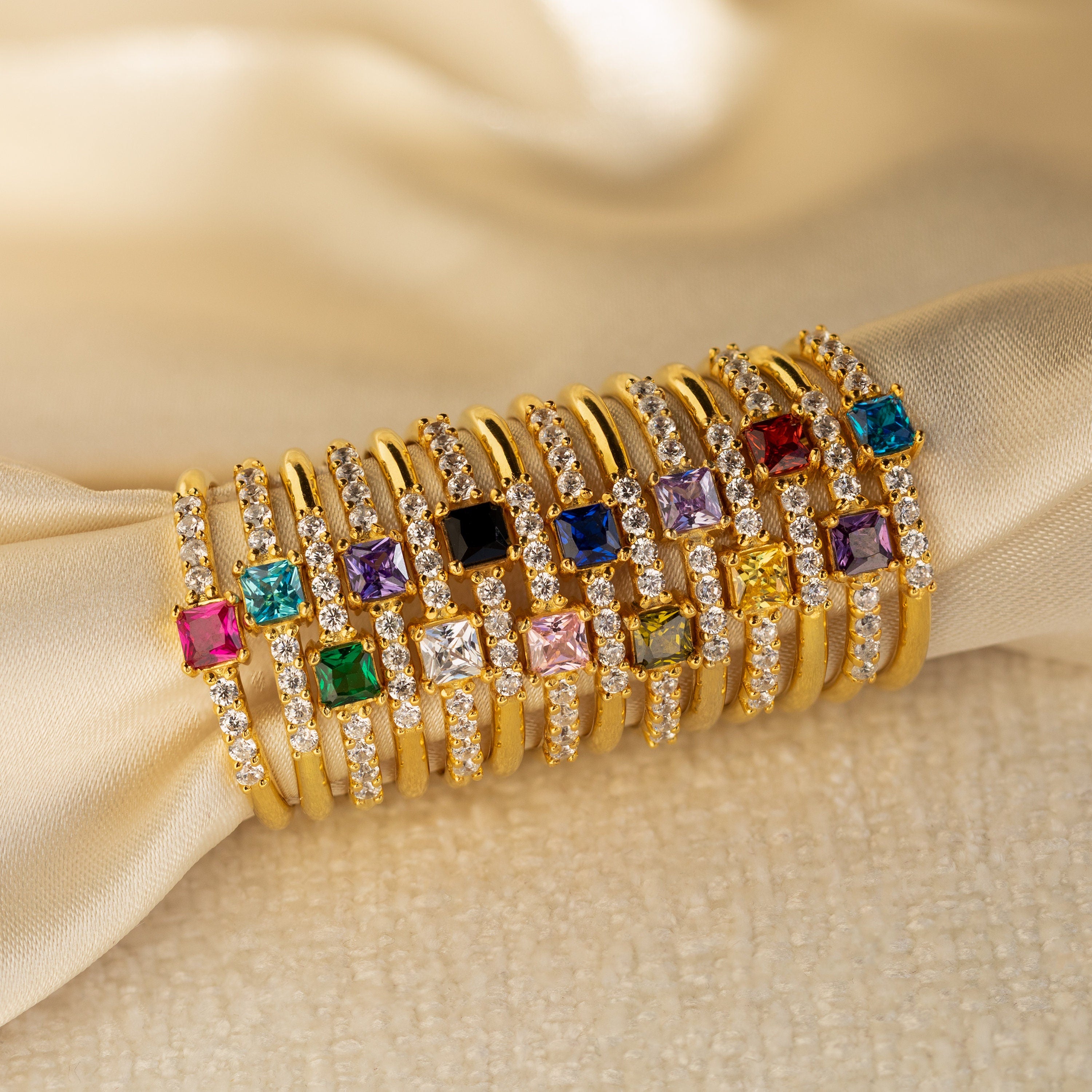 Nine Pave Princess Birthstone Rings with colorful square gemstones and small diamonds—ideal as promise or birthstone rings—are displayed on cream-colored fabric.