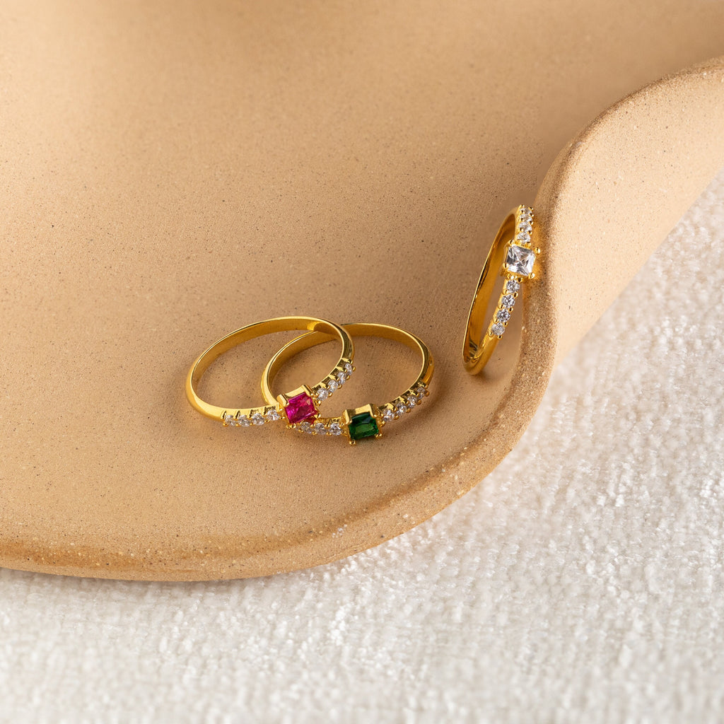 Three Pave Princess Birthstone Rings, each featuring colored gemstones and delicate diamonds, rest elegantly on a beige textured surface.