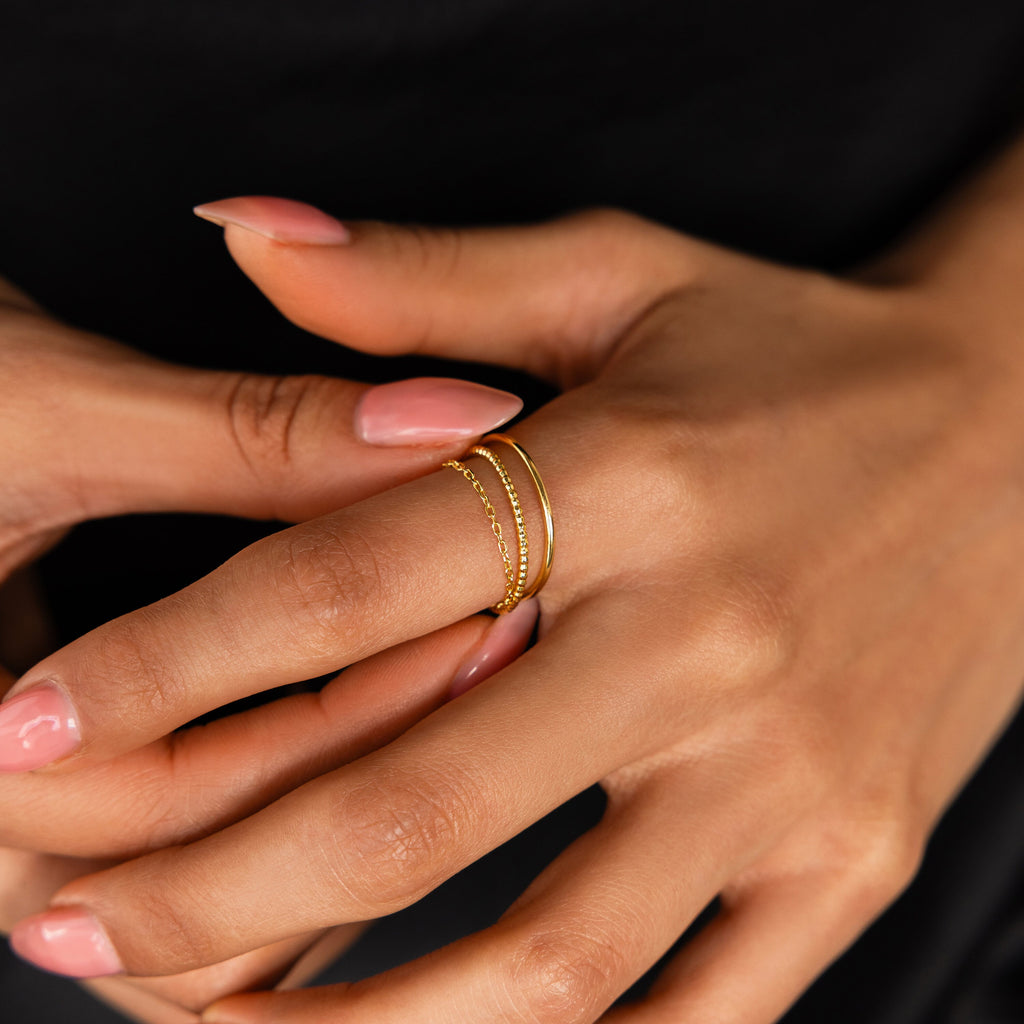 A hand with manicured nails wears the Textured Stacking Ring Set and other dainty rings on the middle finger against a black background.
