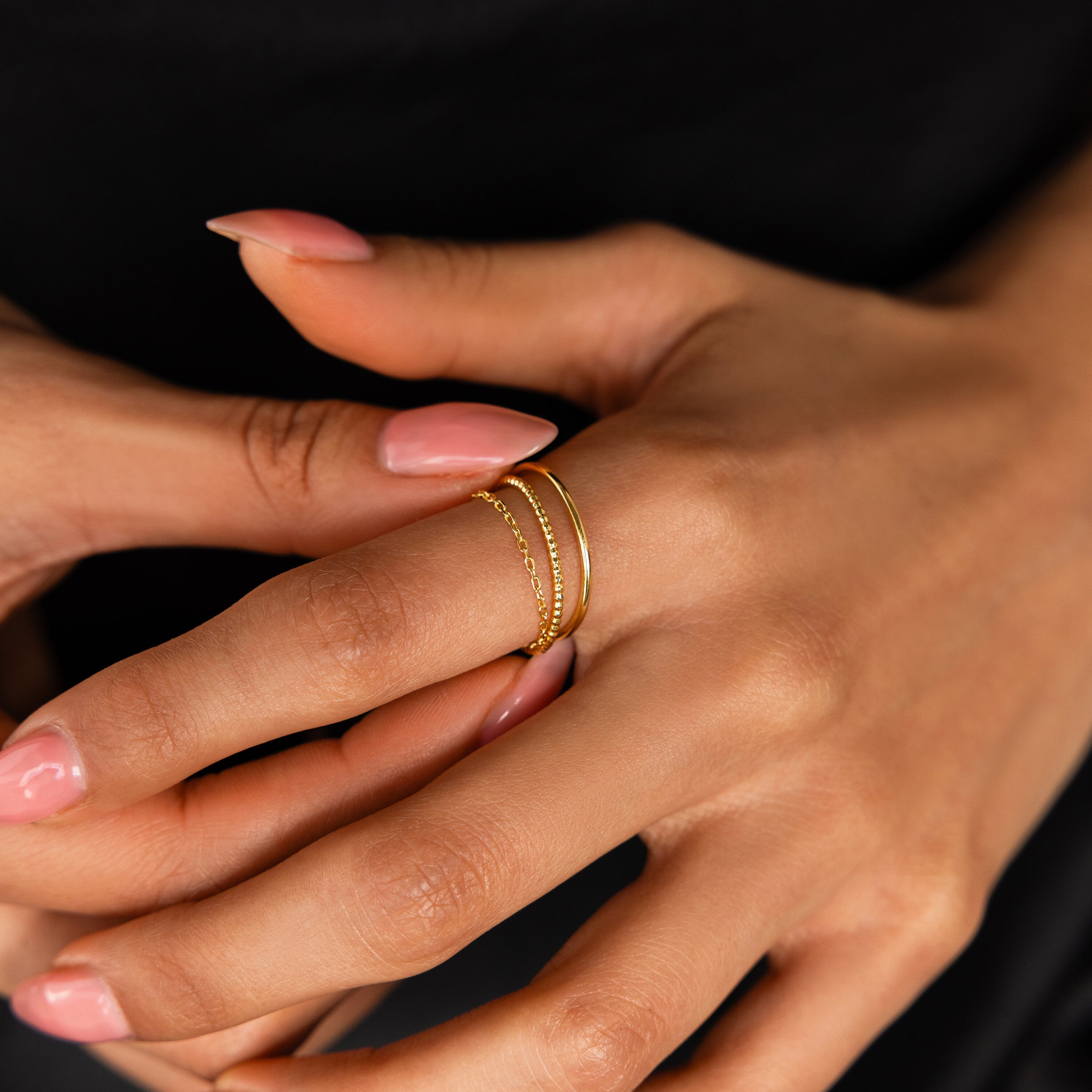 A hand with manicured nails wears the Textured Stacking Ring Set and other dainty rings on the middle finger against a black background.