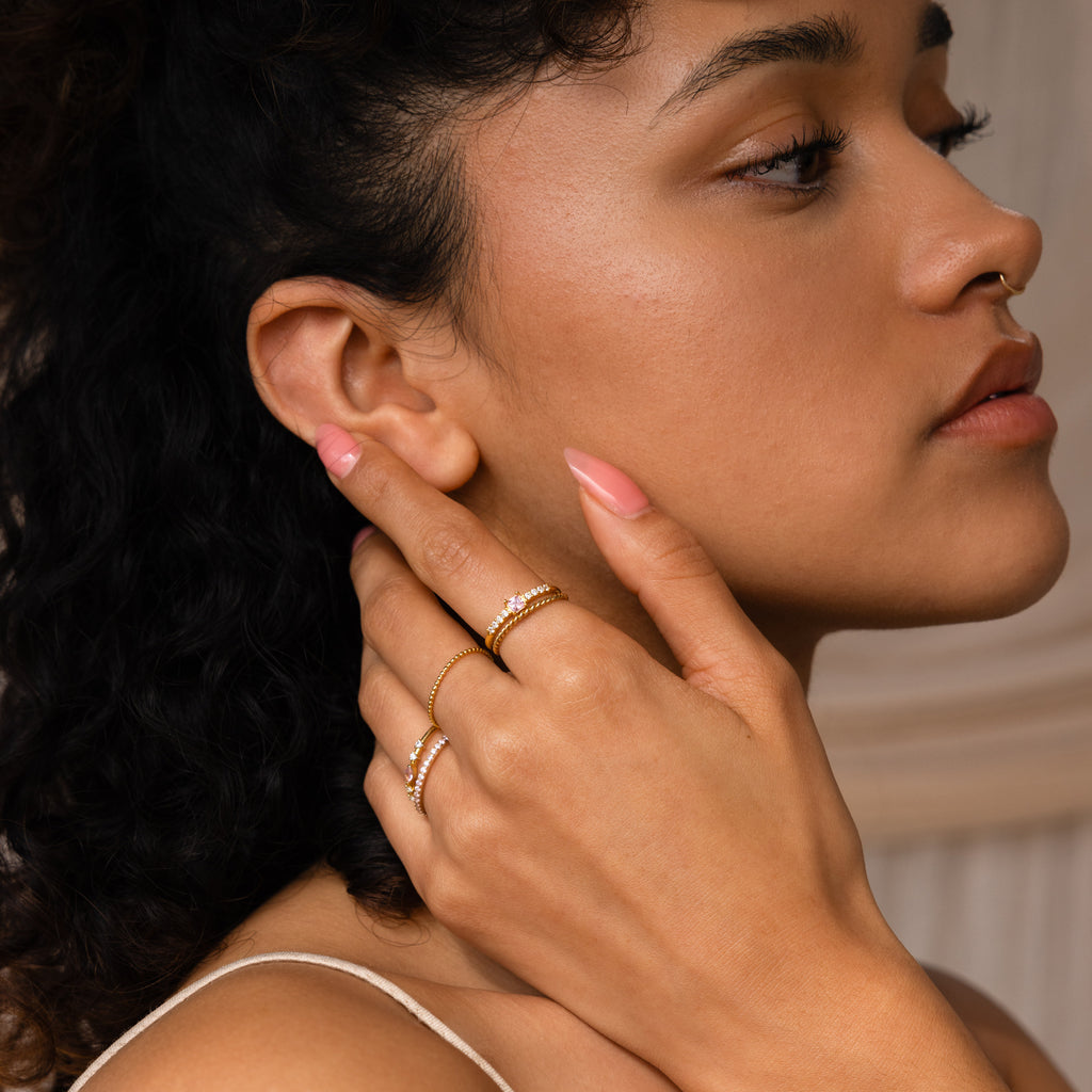 A woman with curly hair and a septum piercing, wearing natural makeup, touches her neck while showcasing the Pave Princess Birthstone Ring.