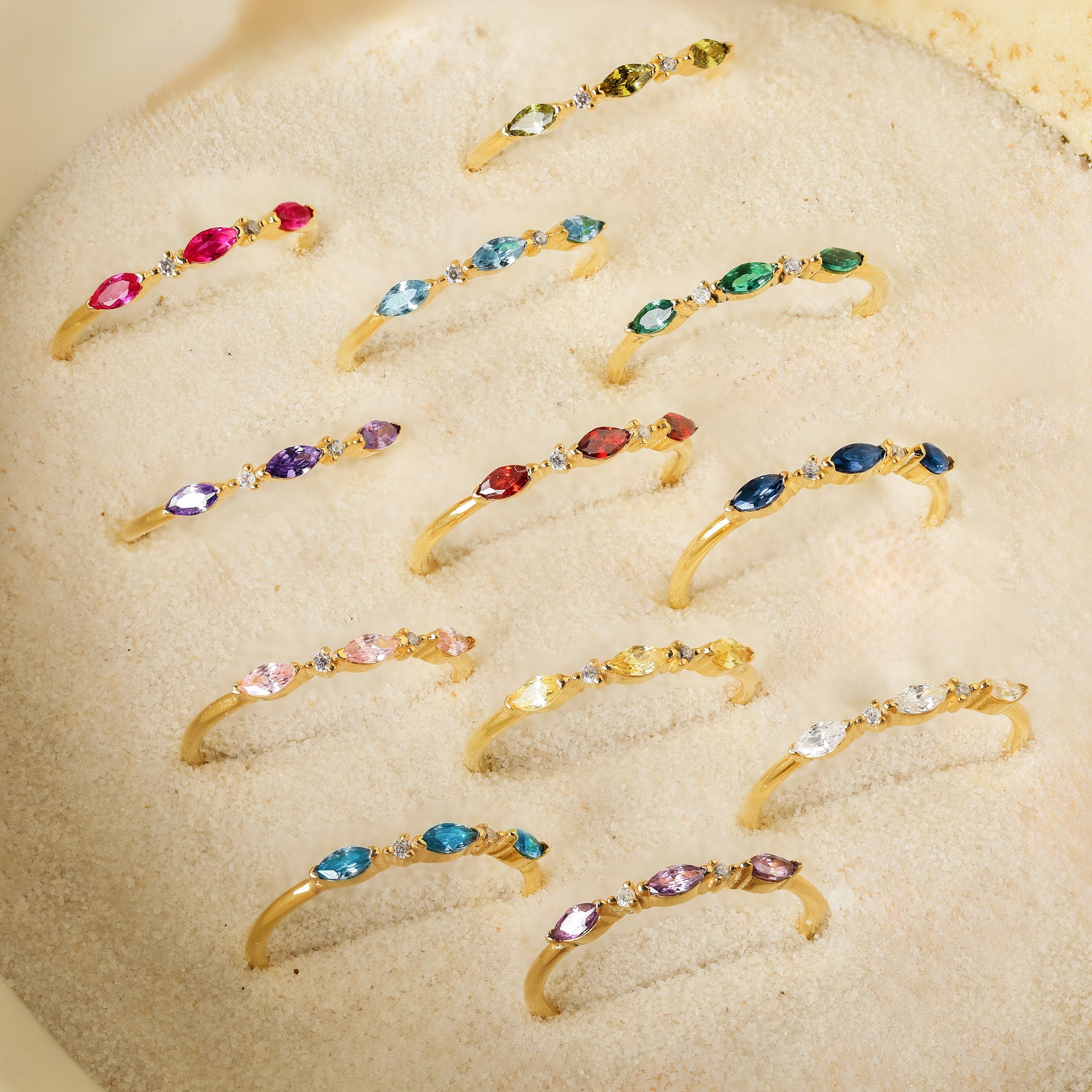 The Triple Marquise Birthstone Ring, featuring vibrant gemstones in a stylish stacking design, is displayed neatly on sandy ground under sunlight.