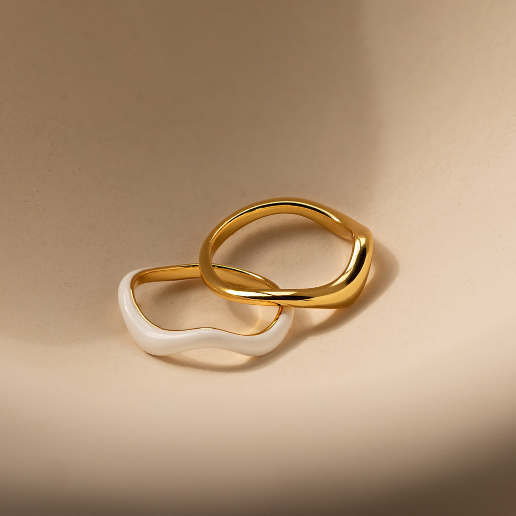 Two wavy rings, one gold and one white, overlap on a beige surface to highlight the Duo Wave Ring Set—a stylish choice for fans of minimalist jewelry.