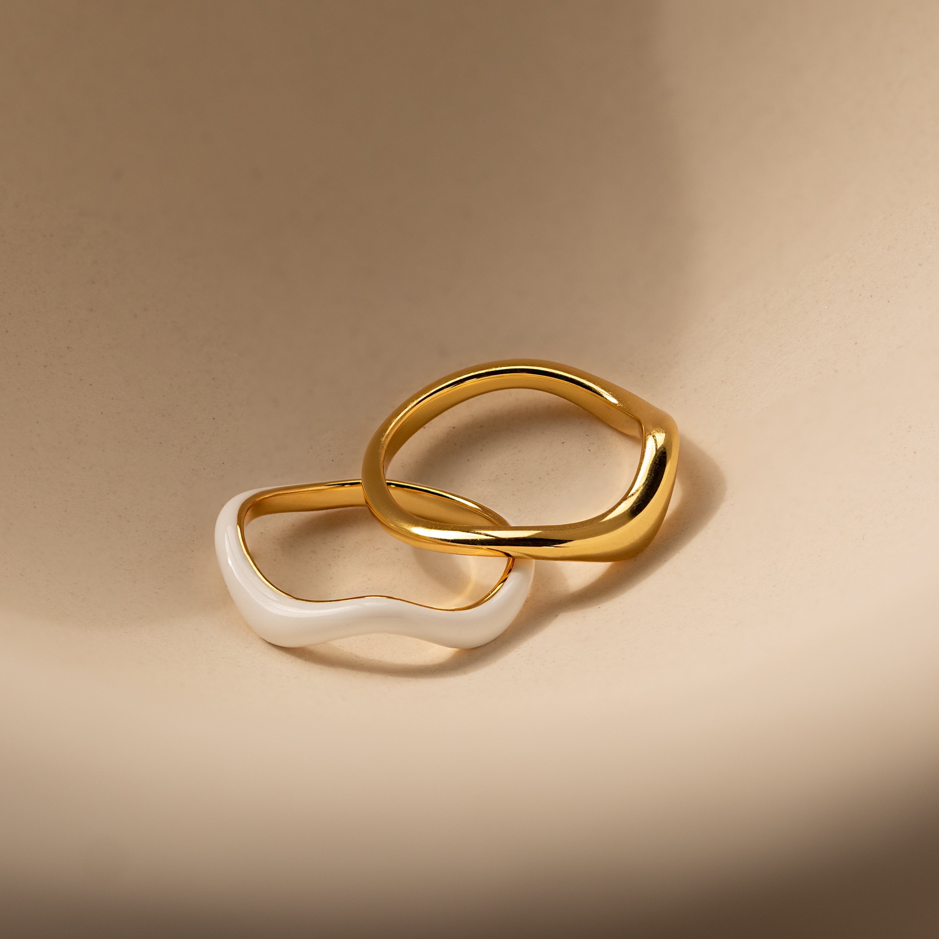 Two wavy rings, one gold and one white, overlap on a beige surface to highlight the Duo Wave Ring Set—a stylish choice for fans of minimalist jewelry.
