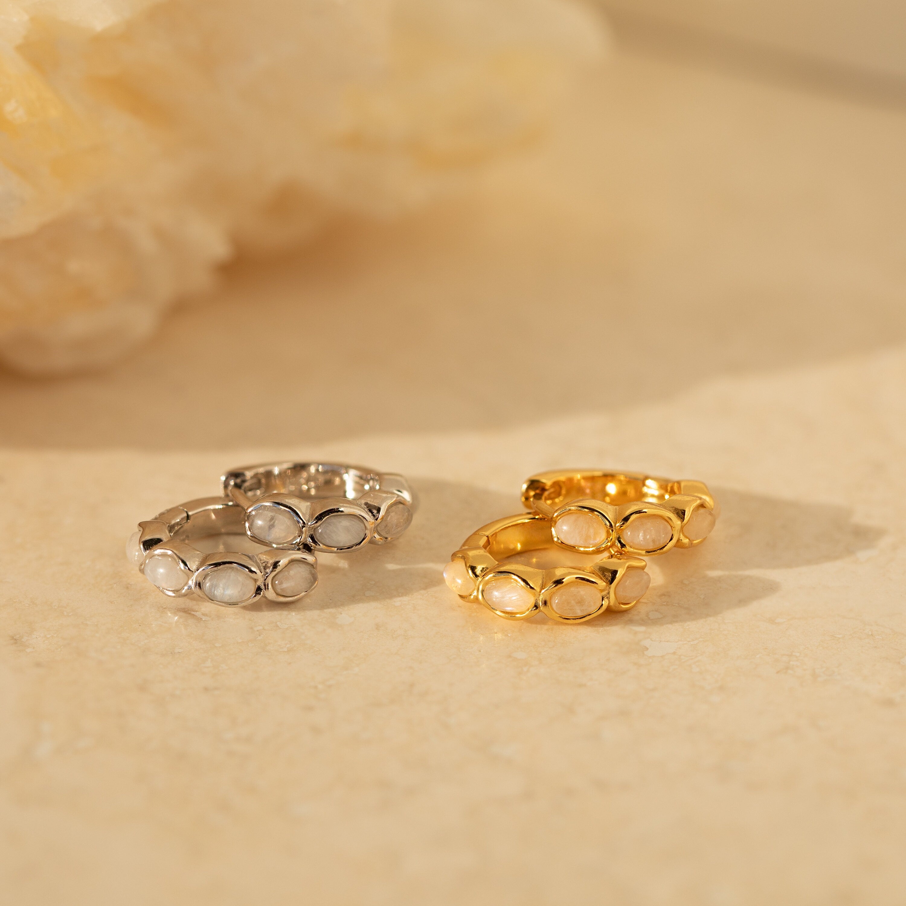 Two pairs of Moonstone Huggies—one in silver, one in gold—rest on a beige surface with a stone backdrop, perfect for fans of minimalist gemstone earrings.