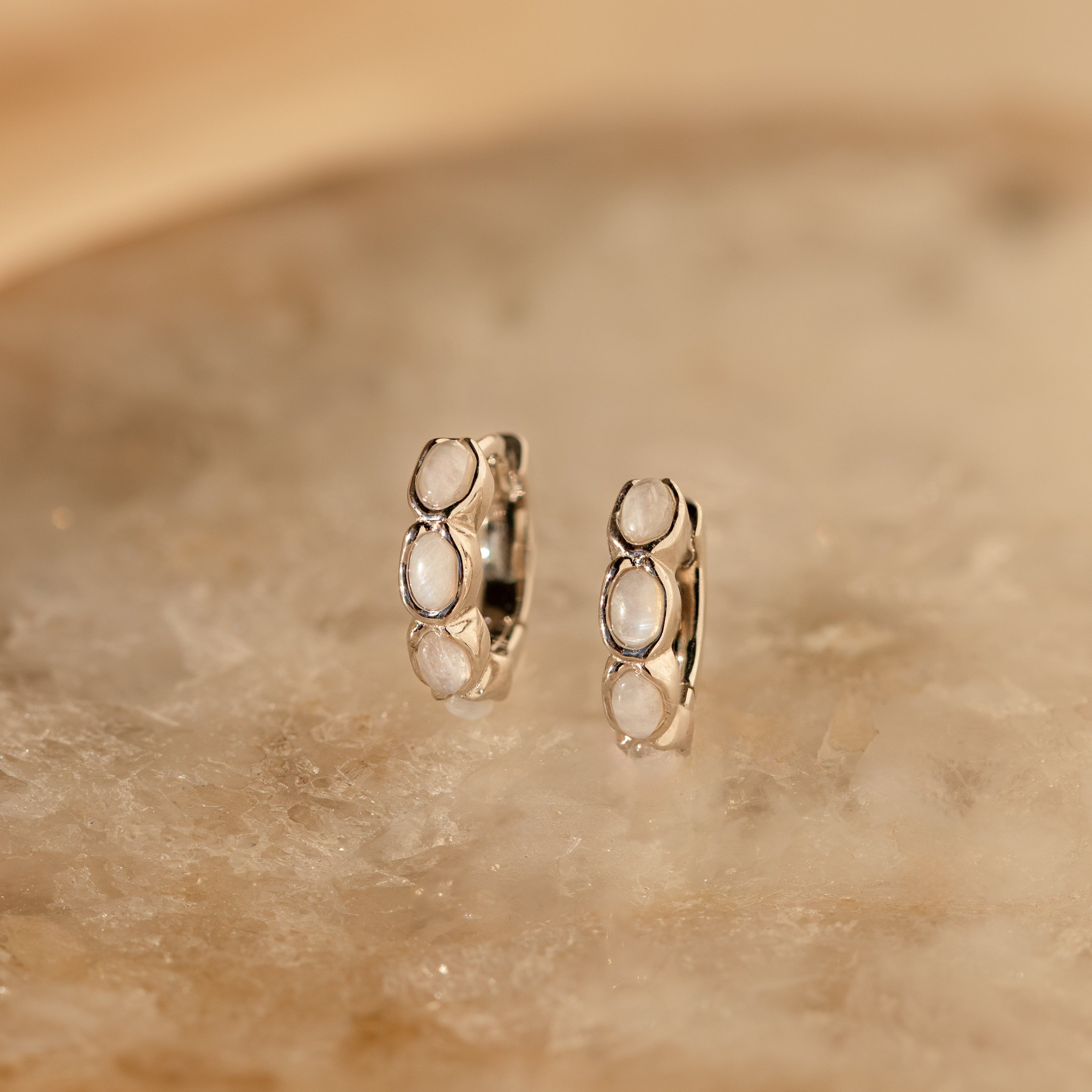The Moonstone Huggies feature three oval white stones set in small silver hoops, showcased on a marbled beige surface—ideal for fans of dainty earrings and minimalist style.