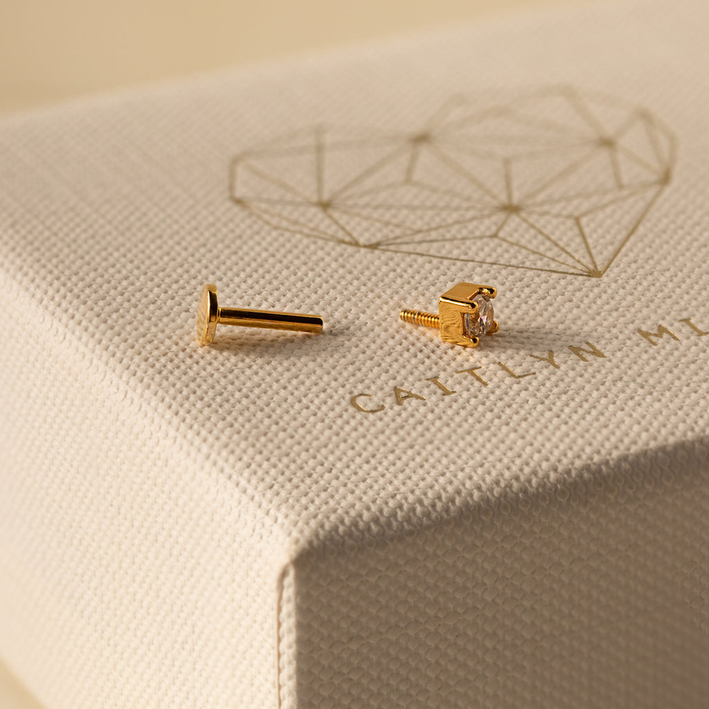 A dainty gold Diamond Flatback Studs Set with clear stones rests on a textured jewelry box featuring a geometric heart design—perfect for lovers of delicate stud earrings.
