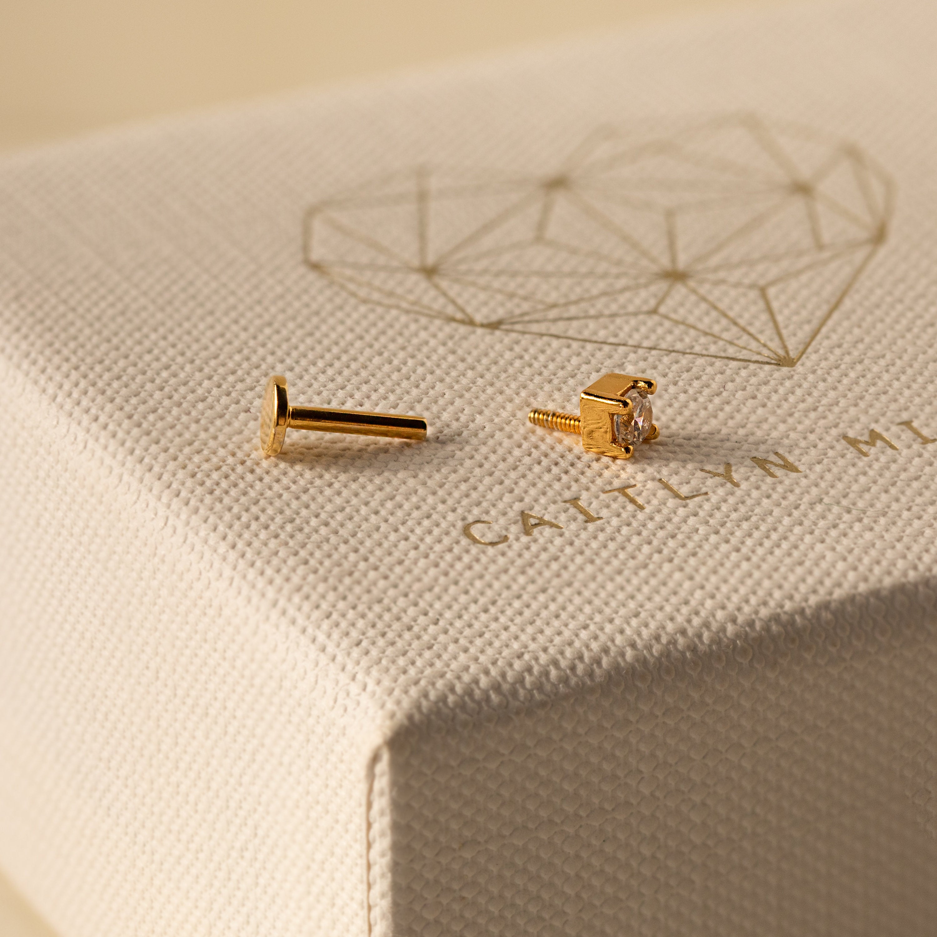 A dainty gold Diamond Flatback Studs Set with clear stones rests on a textured jewelry box featuring a geometric heart design—perfect for lovers of delicate stud earrings.