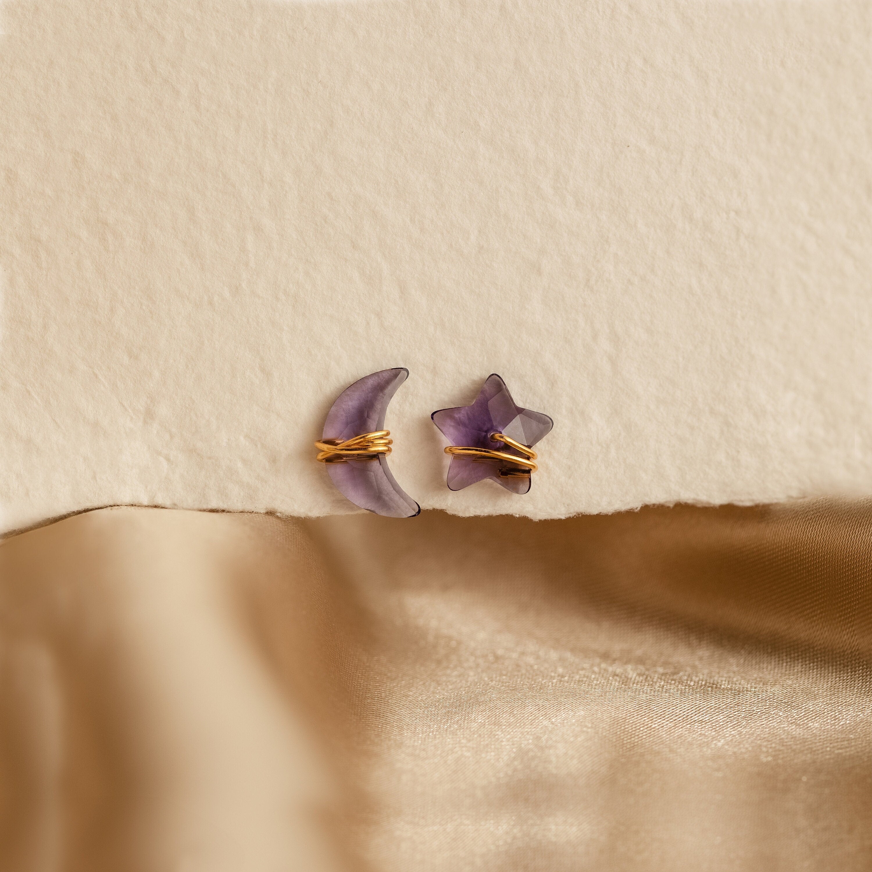 Purple moon and star earrings in gold set against a textured beige background, 