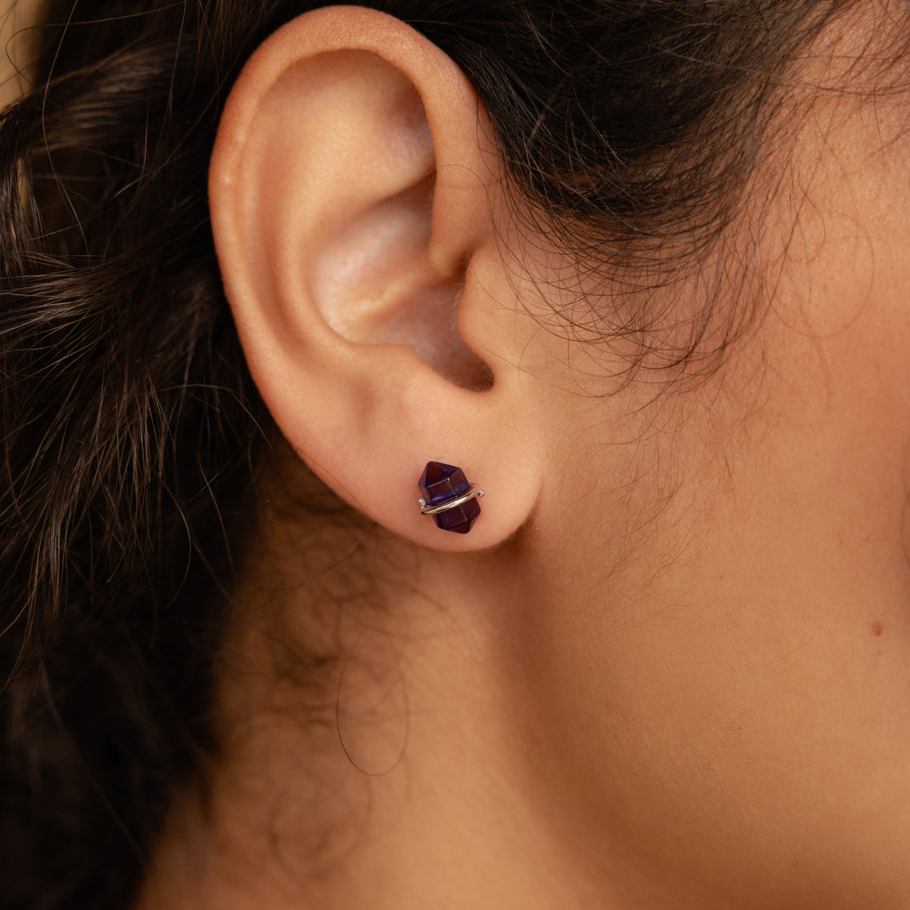 Close-up of a person’s ear wearing Amethyst Herkimer Studs—small, geometric purple earrings crafted from Amethyst Quartz Glass and finished with stylish gold accents.