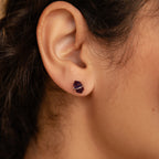 Close-up of a person’s ear wearing Amethyst Herkimer Studs—small, geometric purple earrings crafted from Amethyst Quartz Glass and finished with stylish gold accents.