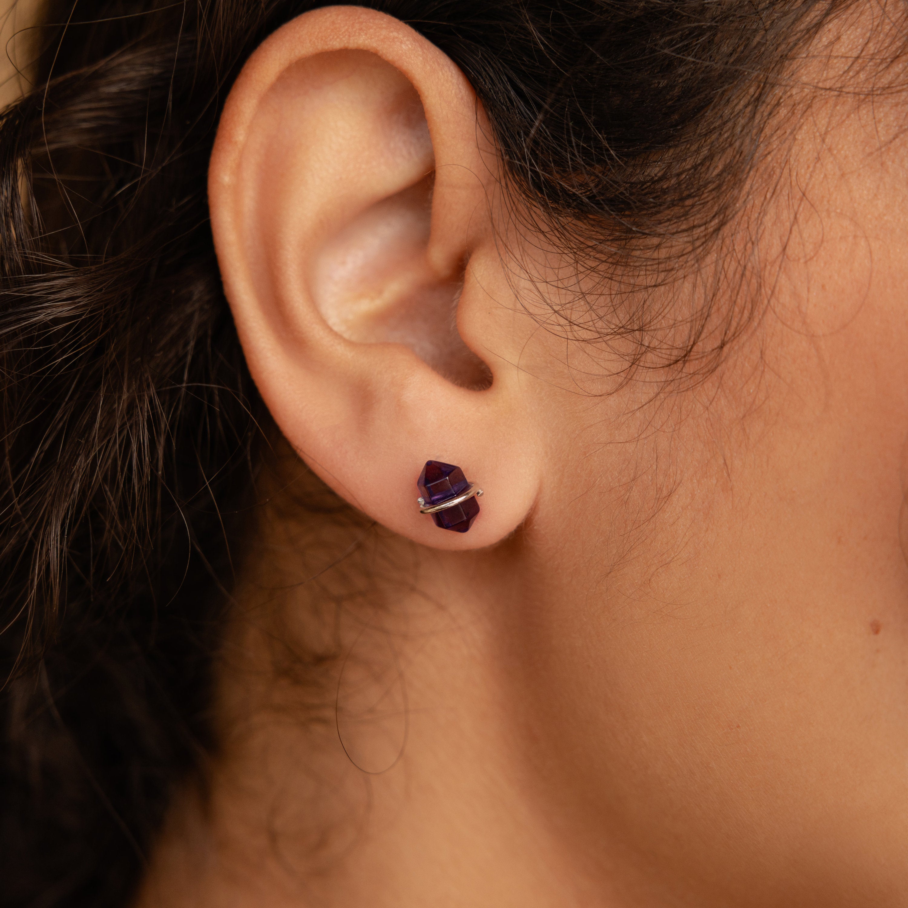 Close-up of a person’s ear wearing Amethyst Herkimer Studs—small, geometric purple earrings crafted from Amethyst Quartz Glass and finished with stylish gold accents.
