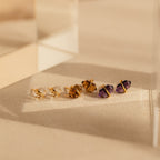 Three pairs of enchanting earrings—clear, brown, and purple Amethyst Herkimer Studs—are beautifully displayed on a beige textured surface.