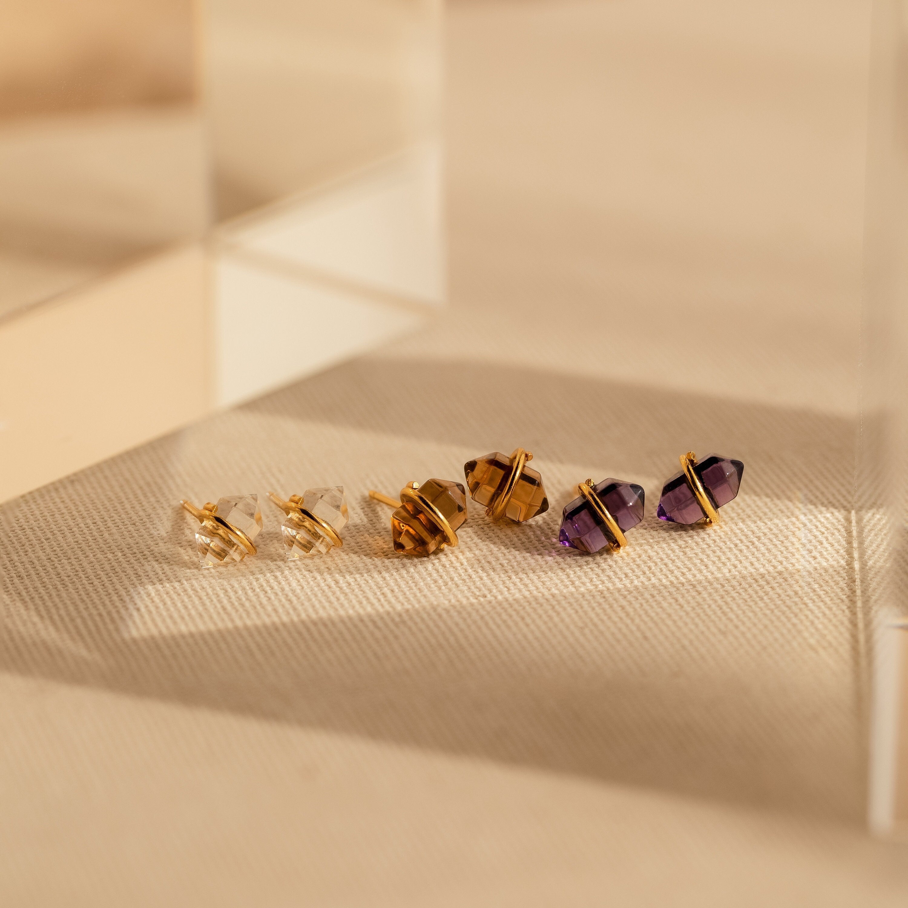 Three pairs of enchanting earrings—clear, brown, and purple Amethyst Herkimer Studs—are beautifully displayed on a beige textured surface.