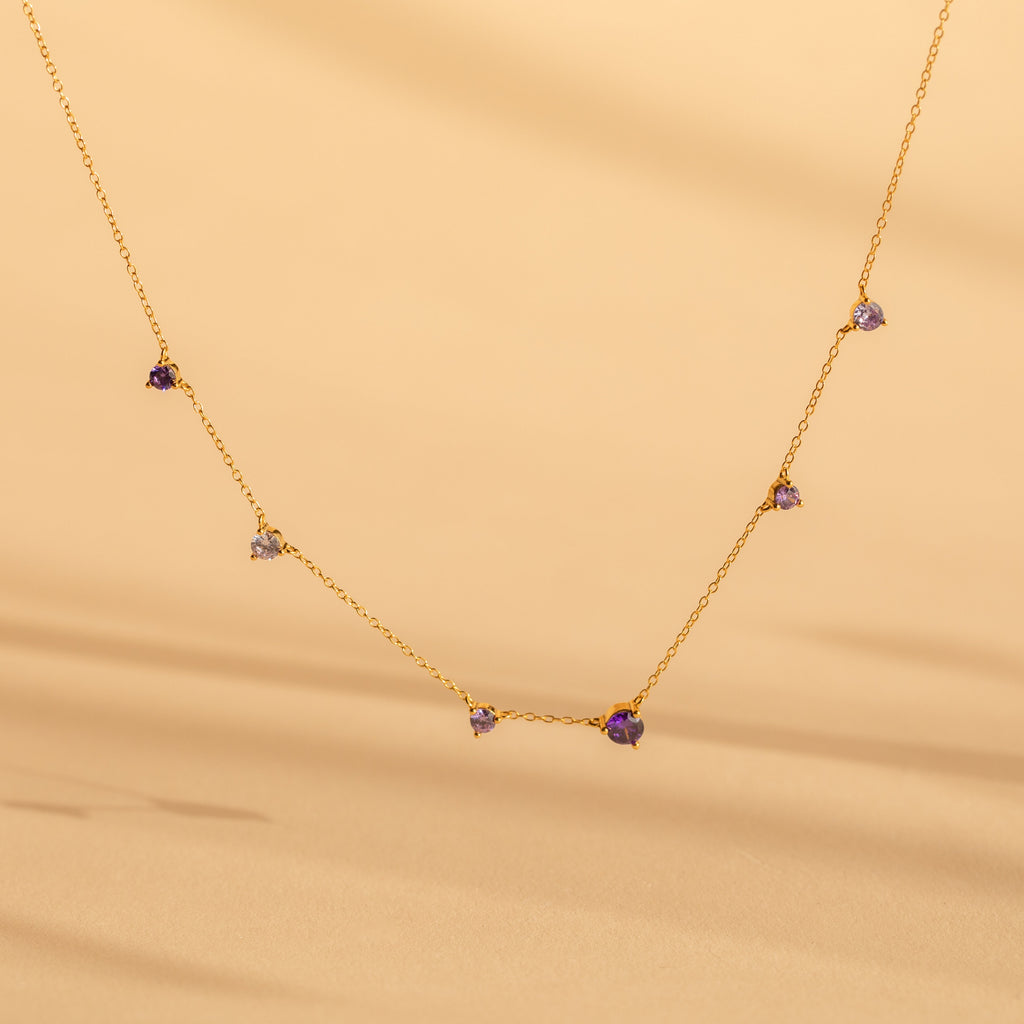 The Amethyst Station Necklace features delicate gold with small purple amethyst stones, elegantly displayed against a soft beige background—a stylish and elegant gift.