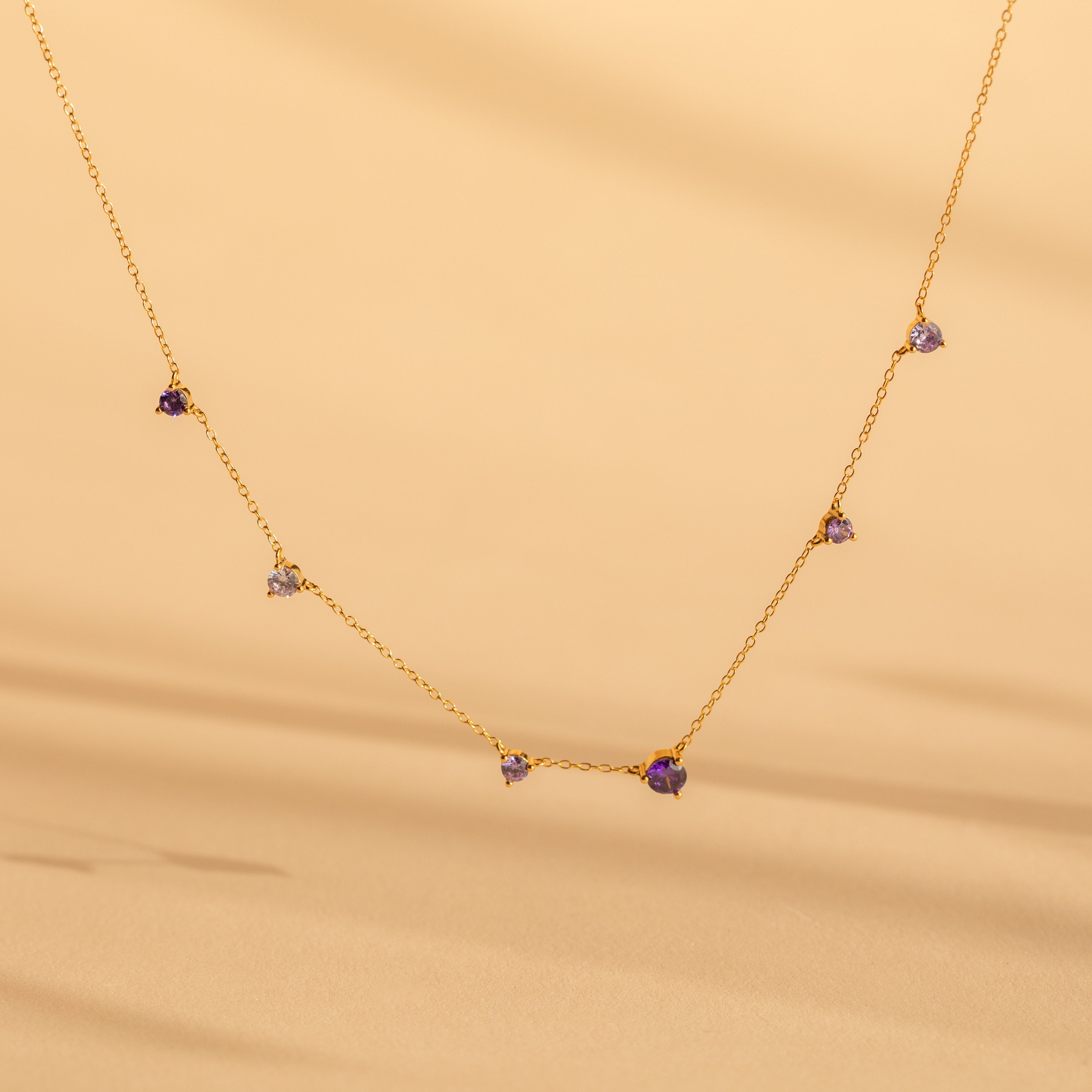 The Amethyst Station Necklace features delicate gold with small purple amethyst stones, elegantly displayed against a soft beige background—a stylish and elegant gift.