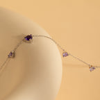 The Amethyst Station Necklace, adorned with purple gemstones, rests elegantly on a smooth beige surface—an ideal, stylish gift for any occasion.