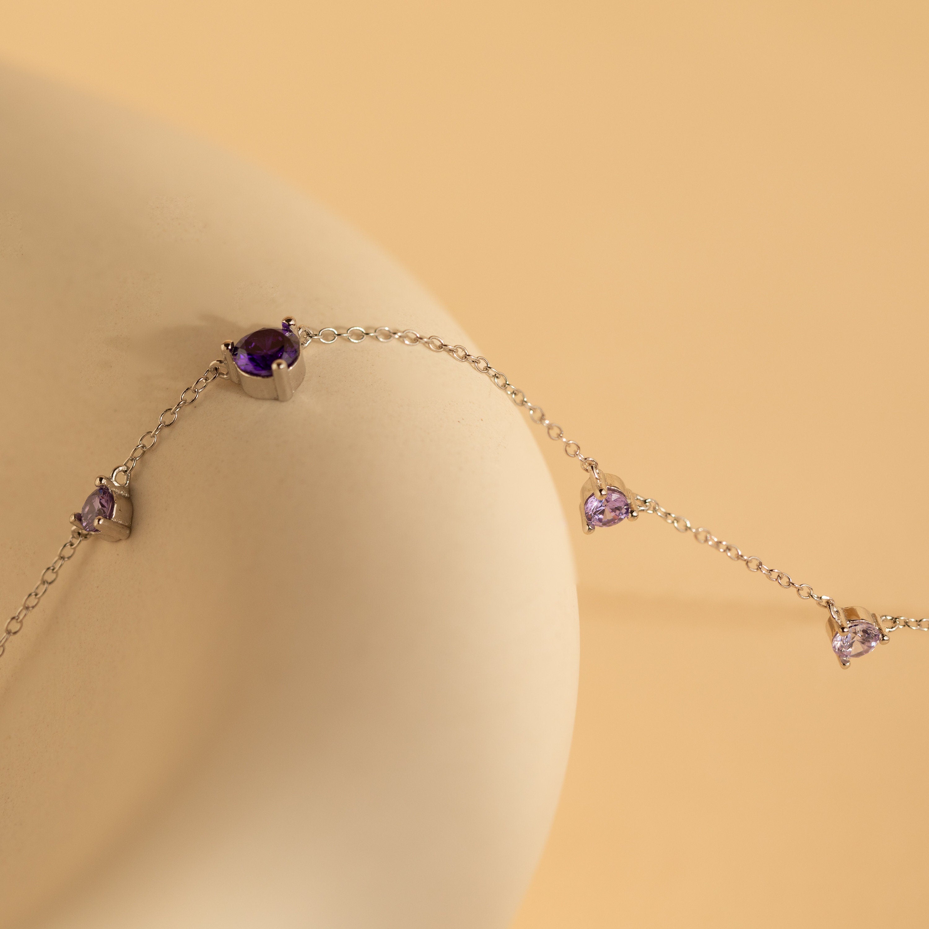 The Amethyst Station Necklace, adorned with purple gemstones, rests elegantly on a smooth beige surface—an ideal, stylish gift for any occasion.