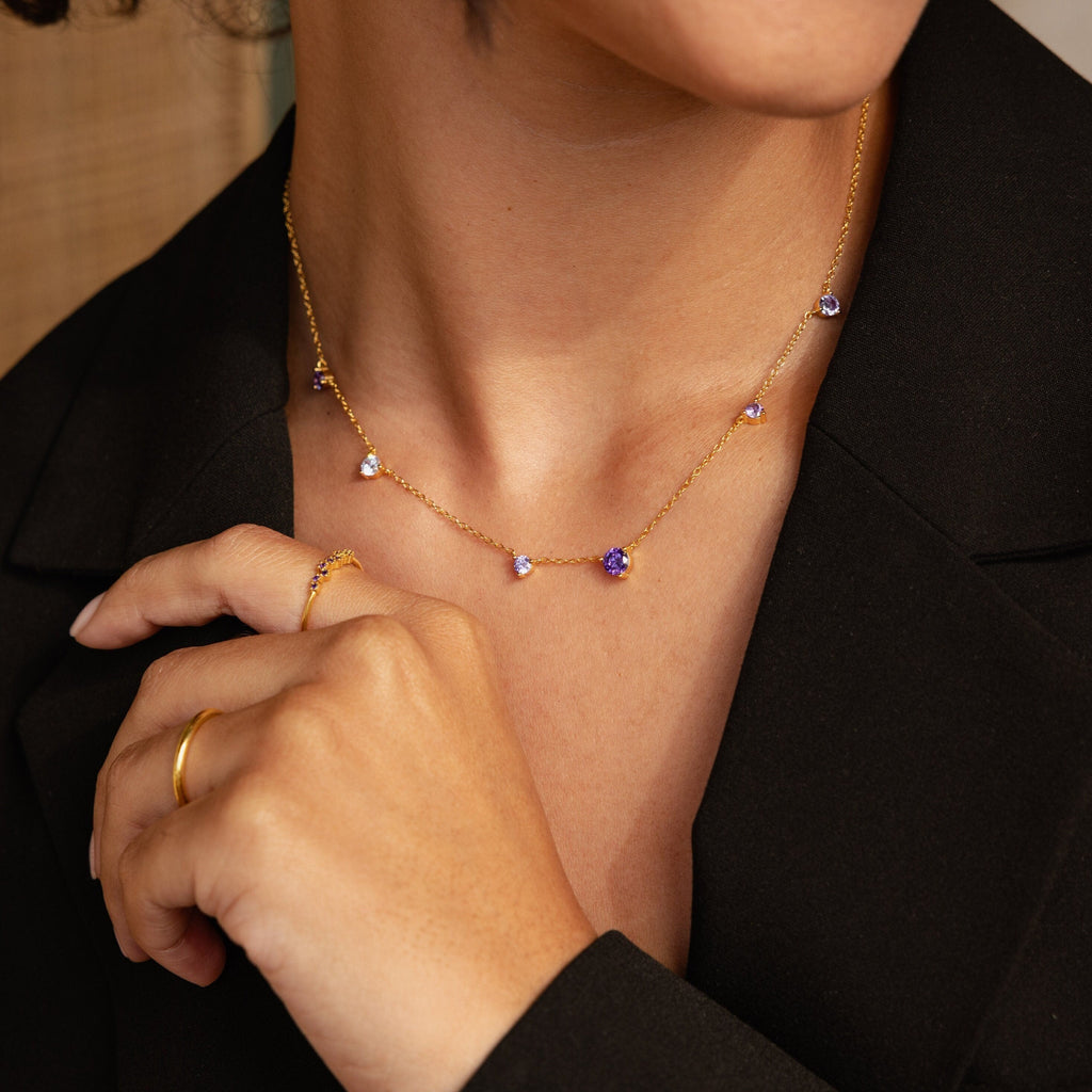 A woman in a black blazer models the Amethyst Station Necklace, adorned with purple amethyst stone charms, and wears a gold ring—a stylish gift.