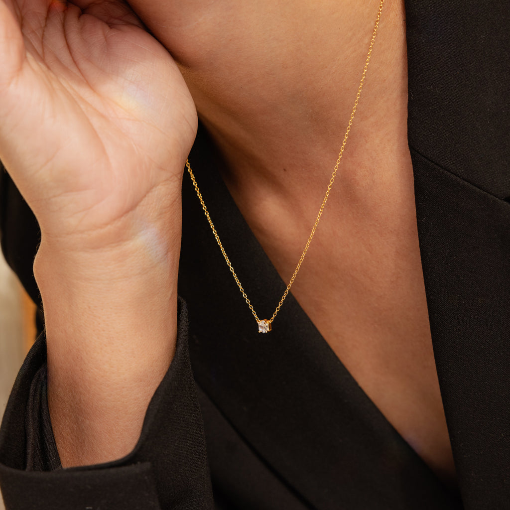 A person wearing the Hexagon Diamond Necklace—a gold geometric piece with a small pendant—paired with a black blazer, hand gently touching their face.