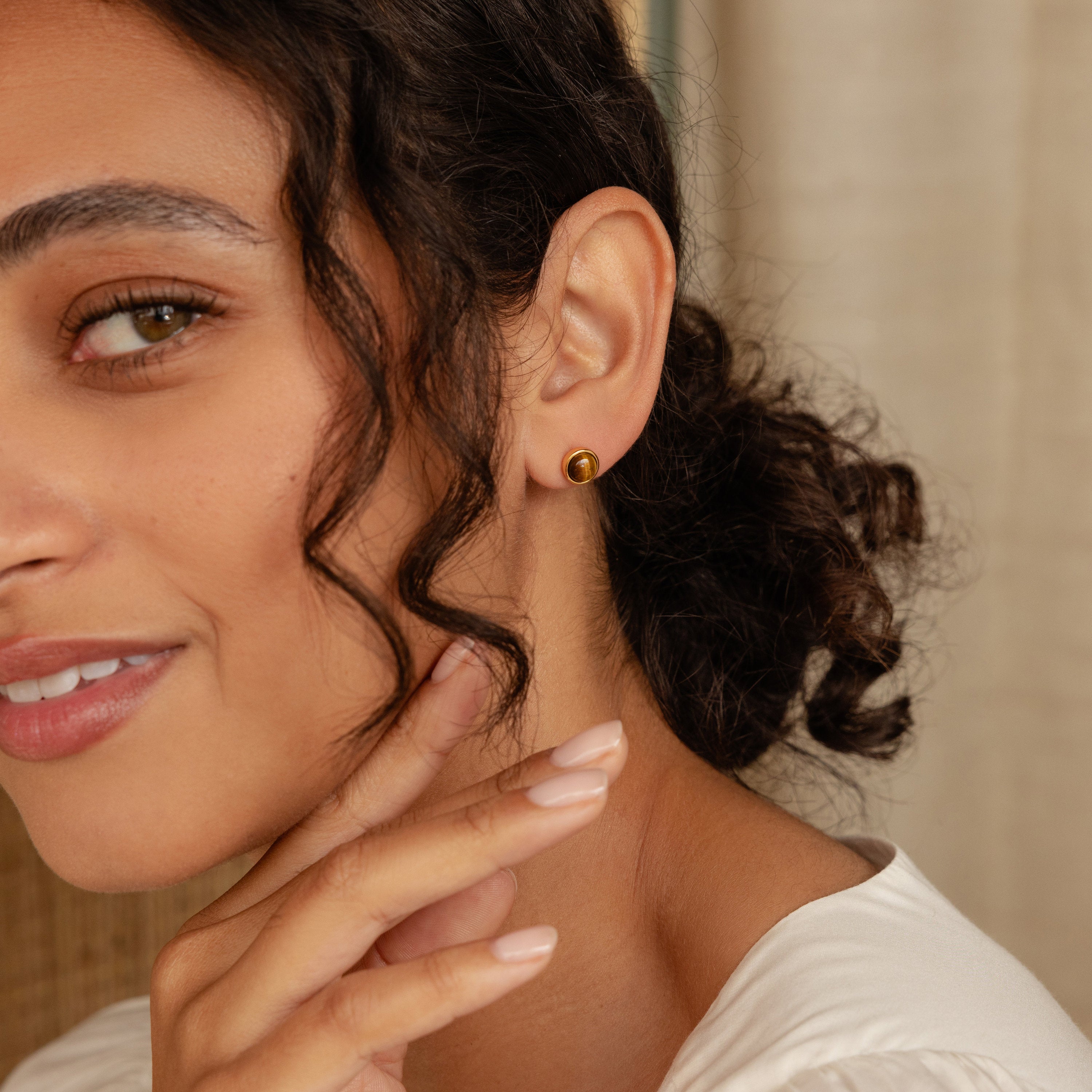 A woman with curly hair wears Manifest Gemstone Studs and touches her face, smiling softly.