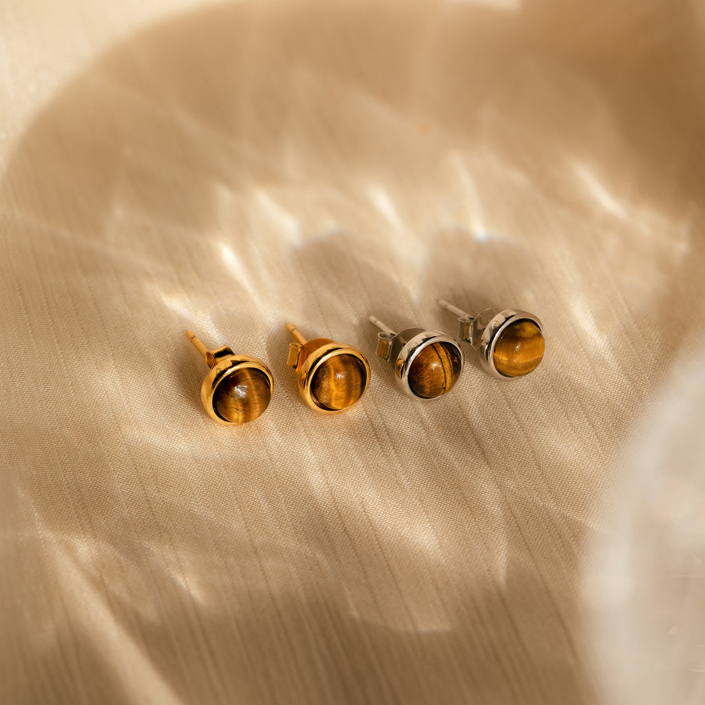 Four round Manifest Gemstone Studs in Tigers Eye—two gold and two silver—are displayed on a beige textured surface under soft light.